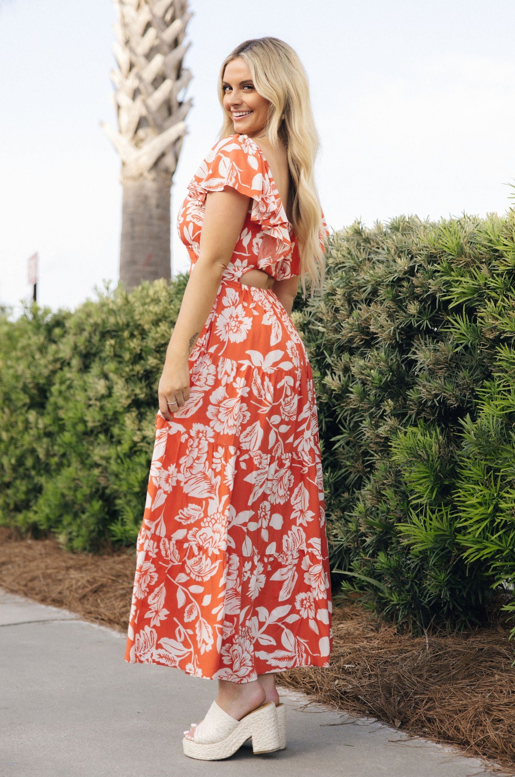 Full body side view of female model wearing the Elena Orange & Cream Floral Midi Dress which features Orange and White Floral Design, Midi Length, Orange Thigh Length Lining, Tiered Body, Smocked Waistband, Sweetheart Neckline, Ruffle Straps and Open Back