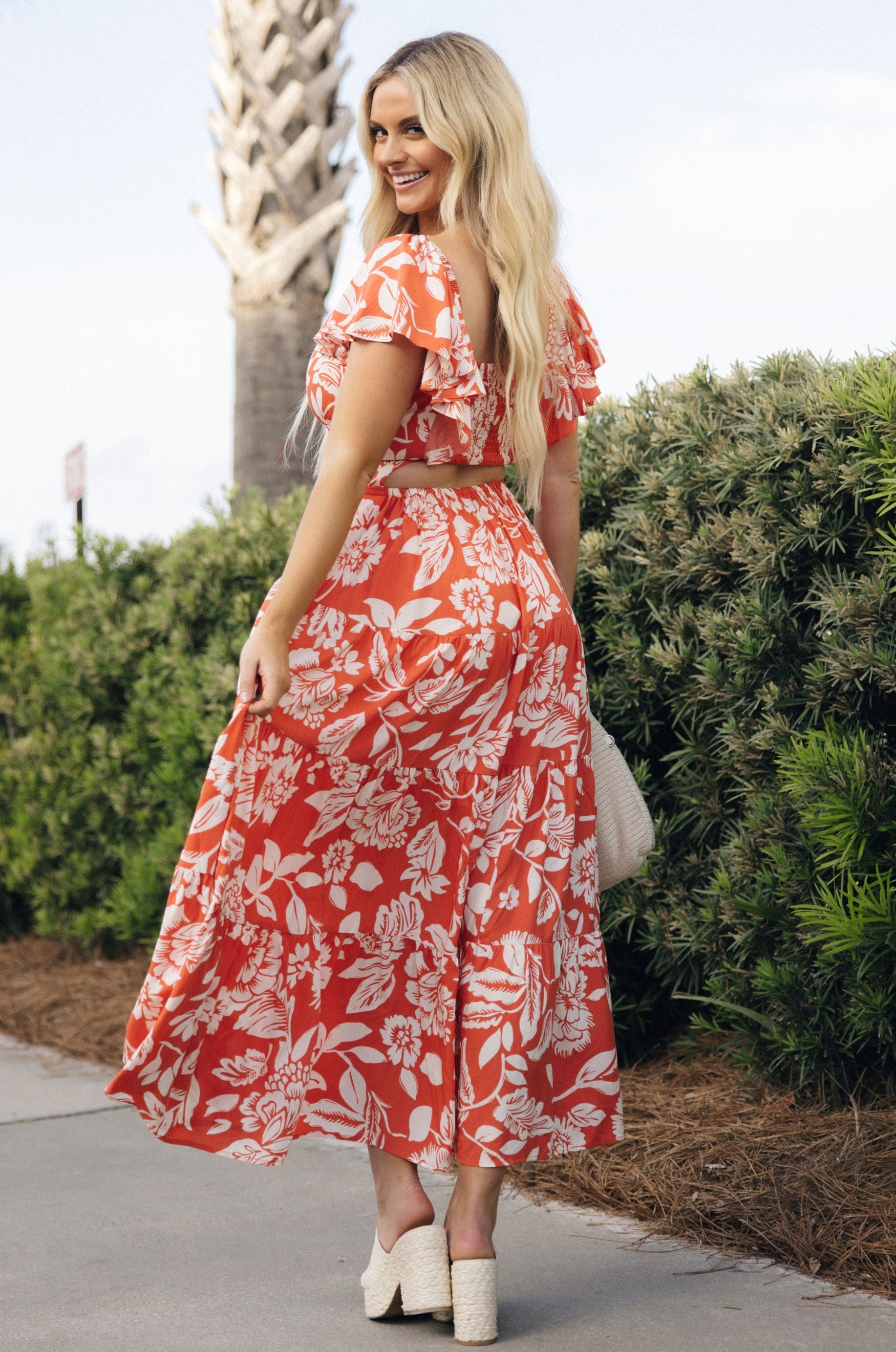 Full body back view of female model wearing the Elena Orange & Cream Floral Midi Dress which features Orange and White Floral Design, Midi Length, Orange Thigh Length Lining, Tiered Body, Smocked Waistband, Sweetheart Neckline, Ruffle Straps and Open Back