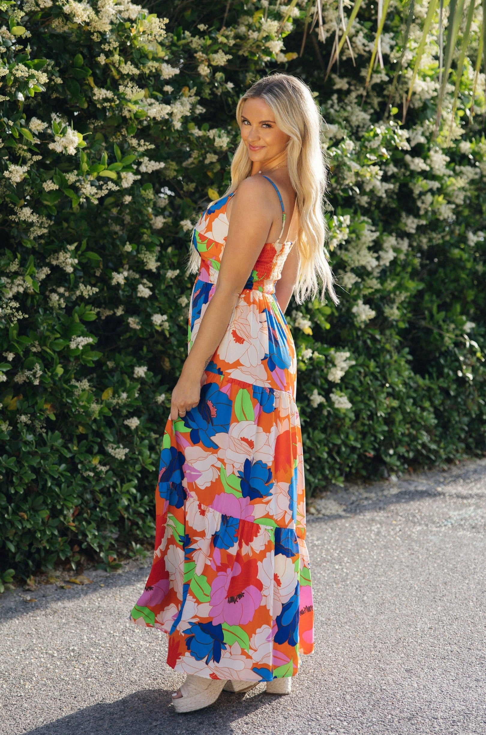 Full body side view of female model wearing the Avianna Orange Floral Maxi Dress that has orange, pink, green, and blue large florals, thin straps, ruffled hem, and ruched bust. 