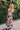 Full body side view of female model wearing the Avianna Orange Floral Maxi Dress that has orange, pink, green, and blue large florals, thin straps, ruffled hem, and ruched bust. 