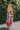 Full body back view of female model wearing the Avianna Orange Floral Maxi Dress that has orange, pink, green, and blue large florals, thin straps, ruffled hem, and ruched bust. 