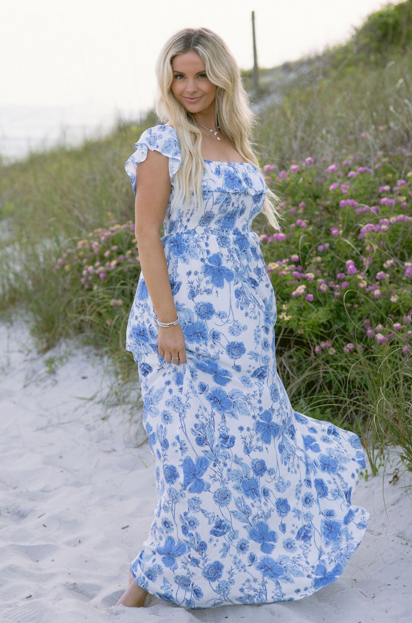 Full body front view of female model wearing the Camila White & Blue Floral Ruffle Maxi Dress which features Blue and White Floral Print, Maxi Skirt, Slit Detail, White Lining, Smocked Upper and Ruffle Straps