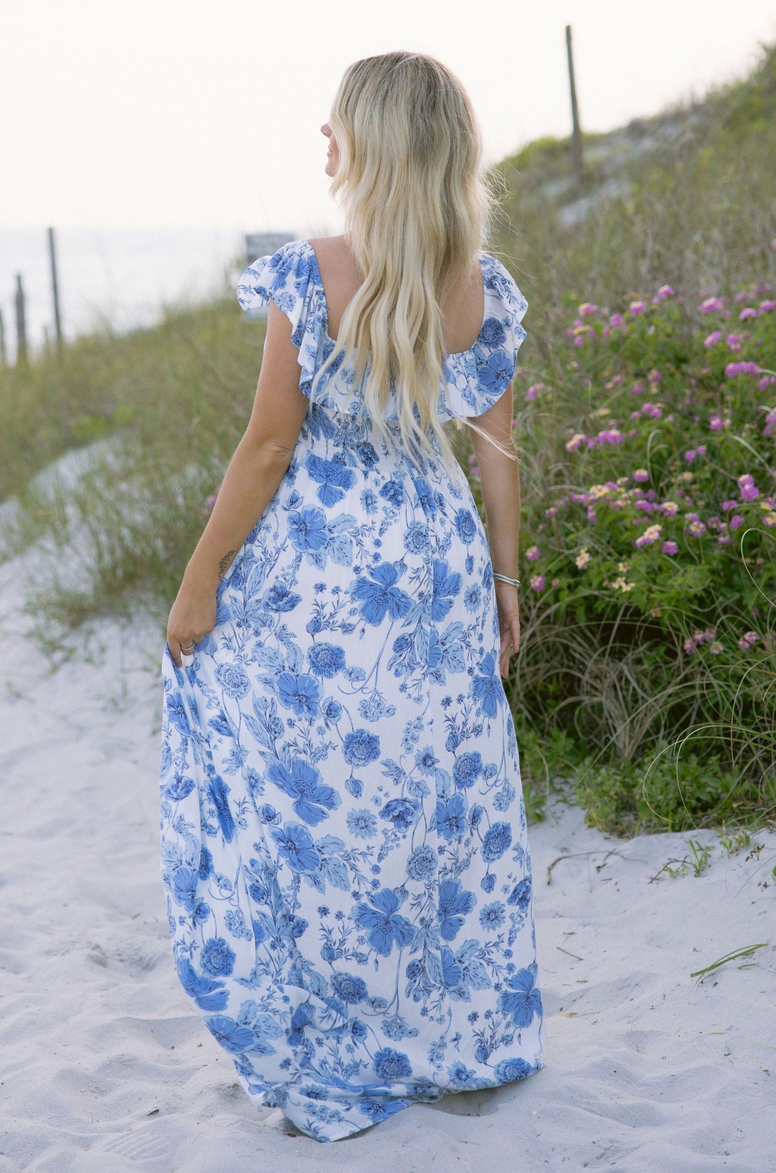 Full body back view of female model wearing the Camila White & Blue Floral Ruffle Maxi Dress which features Blue and White Floral Print, Maxi Skirt, Slit Detail, White Lining, Smocked Upper and Ruffle Straps