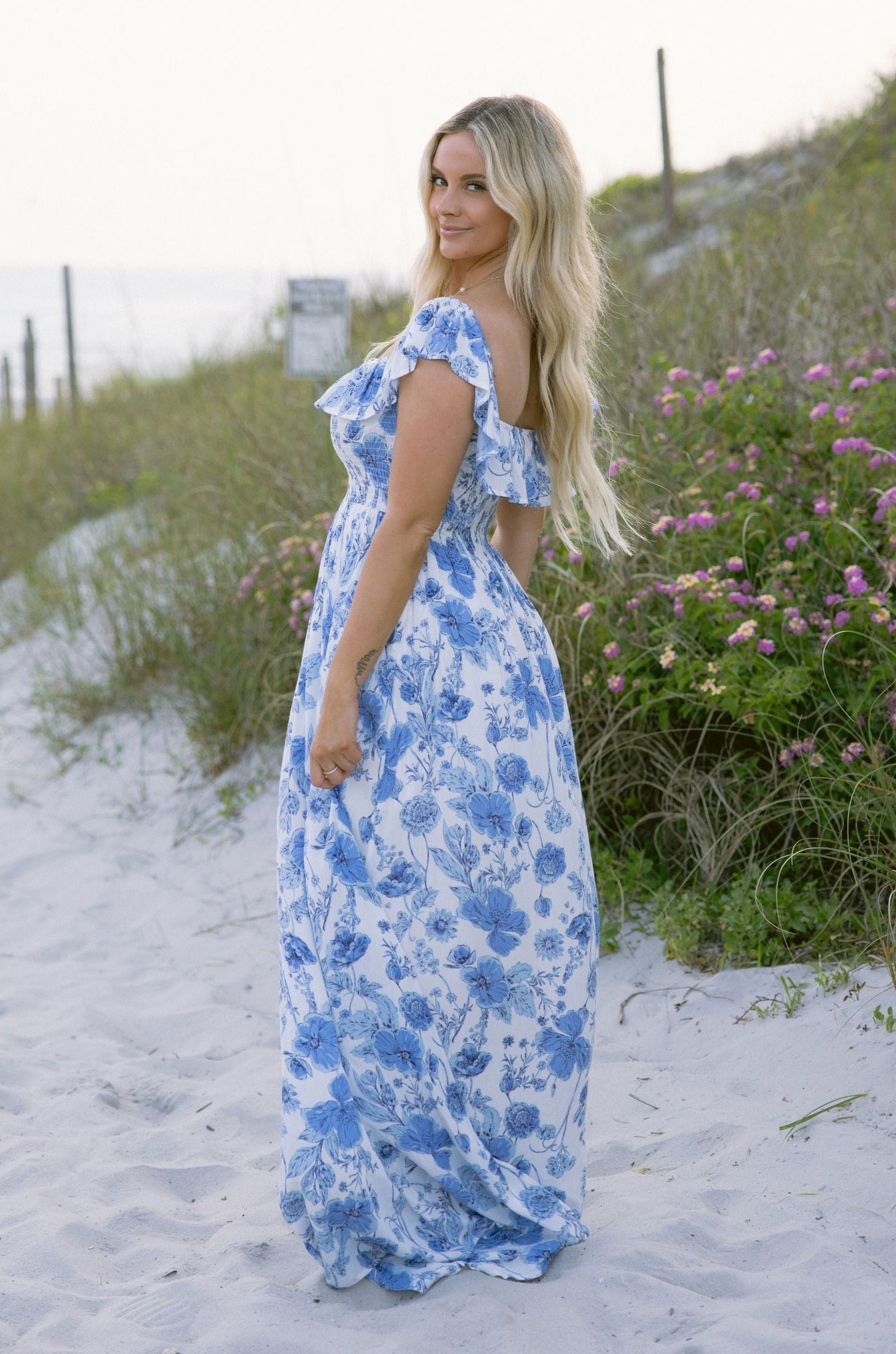 Full body side view of female model wearing the Camila White & Blue Floral Ruffle Maxi Dress which features Blue and White Floral Print, Maxi Skirt, Slit Detail, White Lining, Smocked Upper and Ruffle Straps