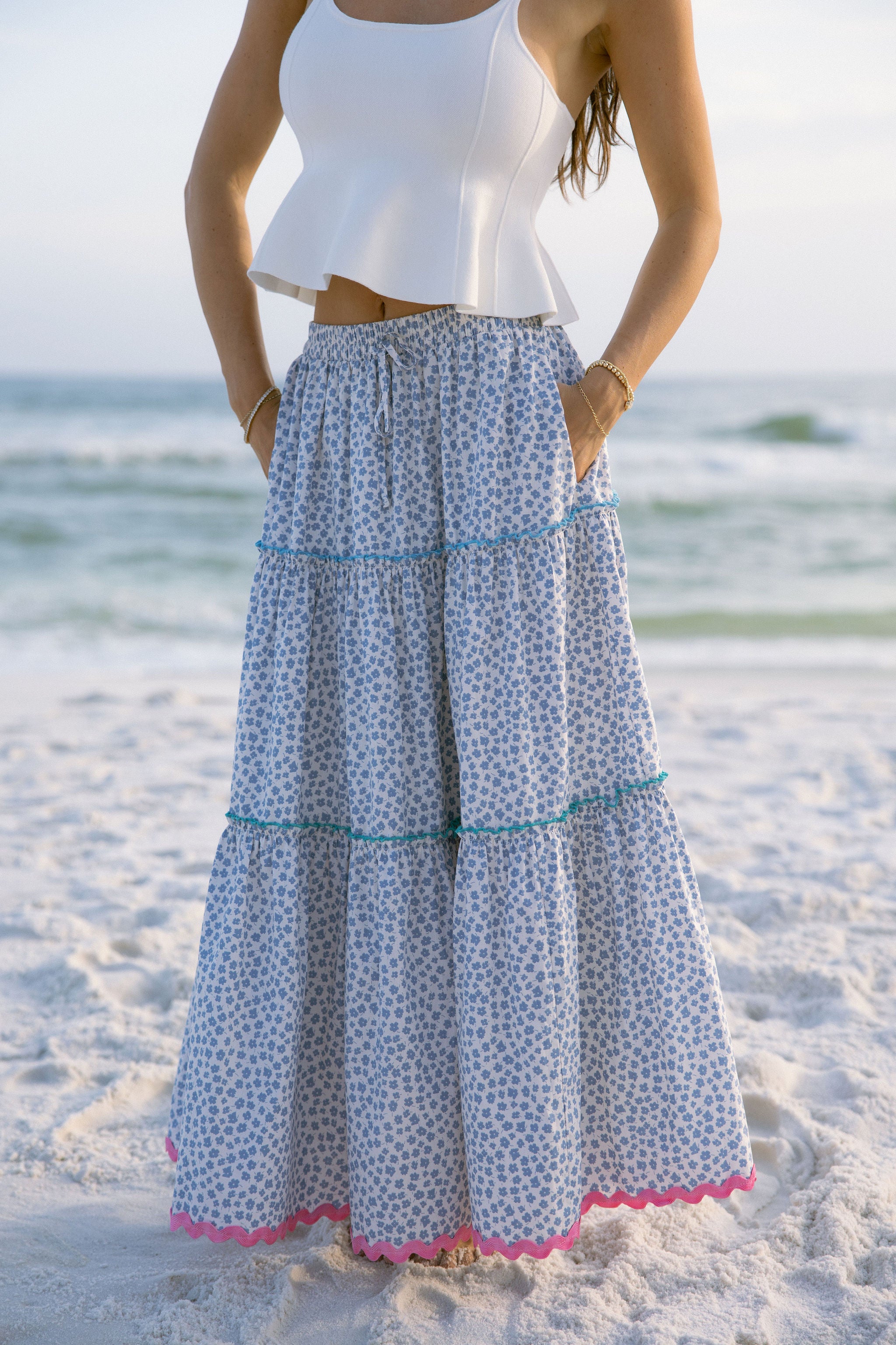 Lower body front view of female model wearing the Marisol Tiered Floral Maxi Skirt in Blue, that has blue and white floral fabric, colorful tiered ruffle trim, and an elastic waist. Worn with white tank top.