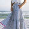 Lower body side view of female model wearing the Marisol Tiered Floral Maxi Skirt in Blue, that has blue and white floral fabric, colorful tiered ruffle trim, and an elastic waist. Worn with white tank top.