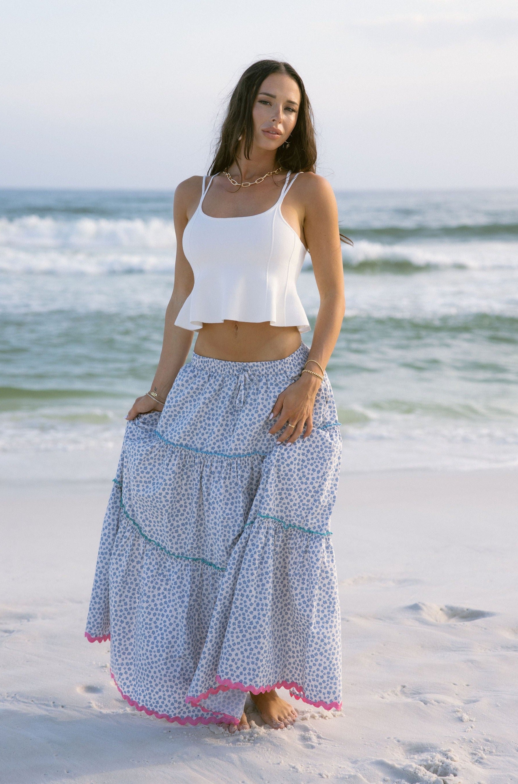 Full body front view of female model wearing the Marisol Tiered Floral Maxi Skirt in Blue, that has blue and white floral fabric, colorful tiered ruffle trim, and an elastic waist. Worn with white tank top.