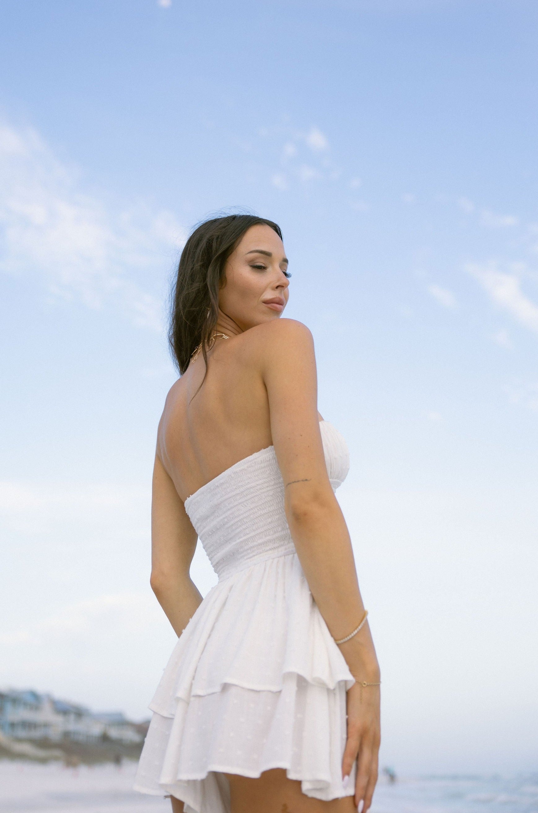 Upper body side view of female model wearing the Jemma White Swiss Dot Romper that has white swiss dot fabric, a strapless neck, and tiered shorts.