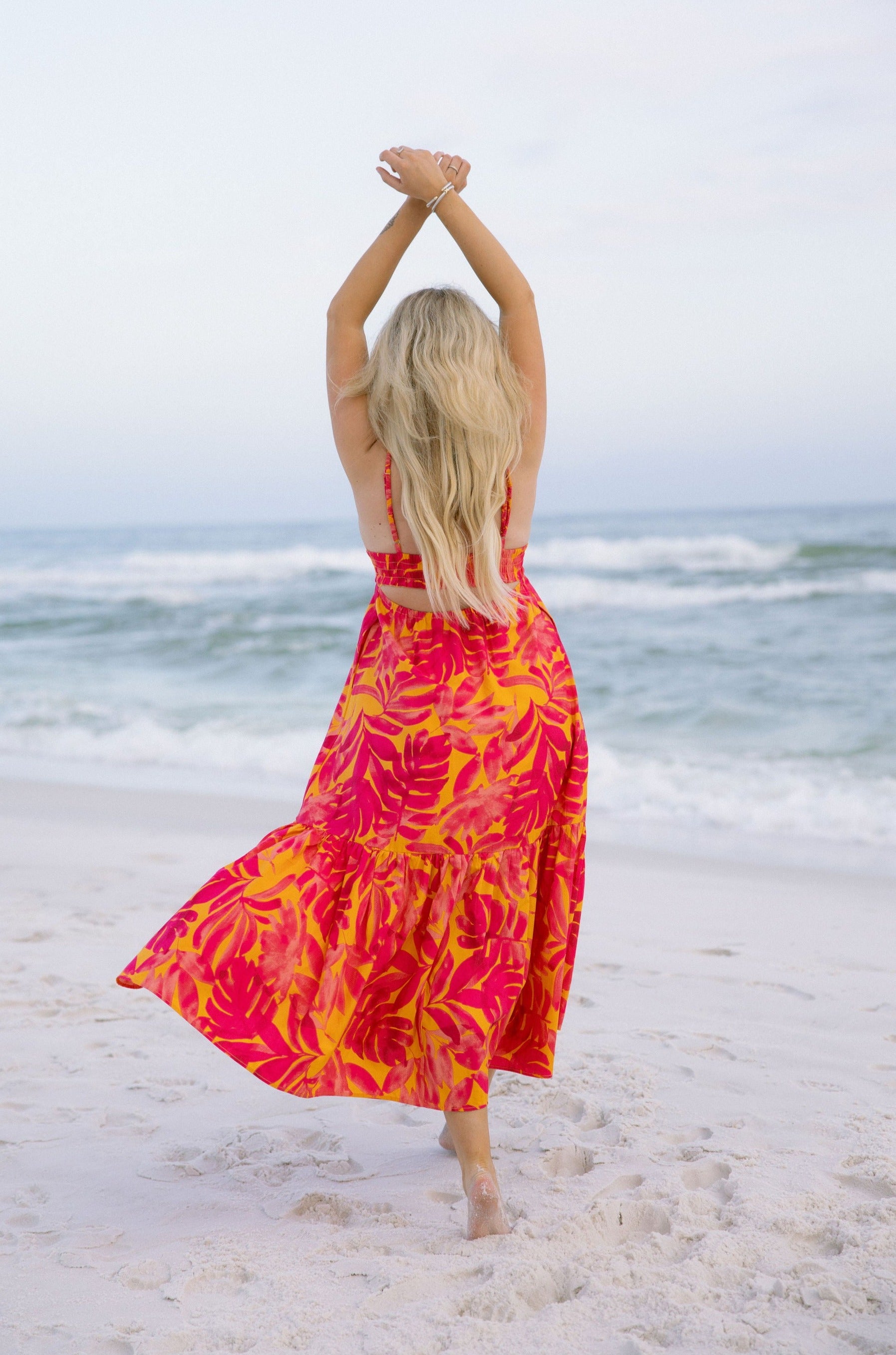 Full body back view of female model wearing the Melina Orange & Pink Floral Maxi Dress that has bright orange and pink floral pattern, adjustable straps, and an open back. Model has arms in air.
