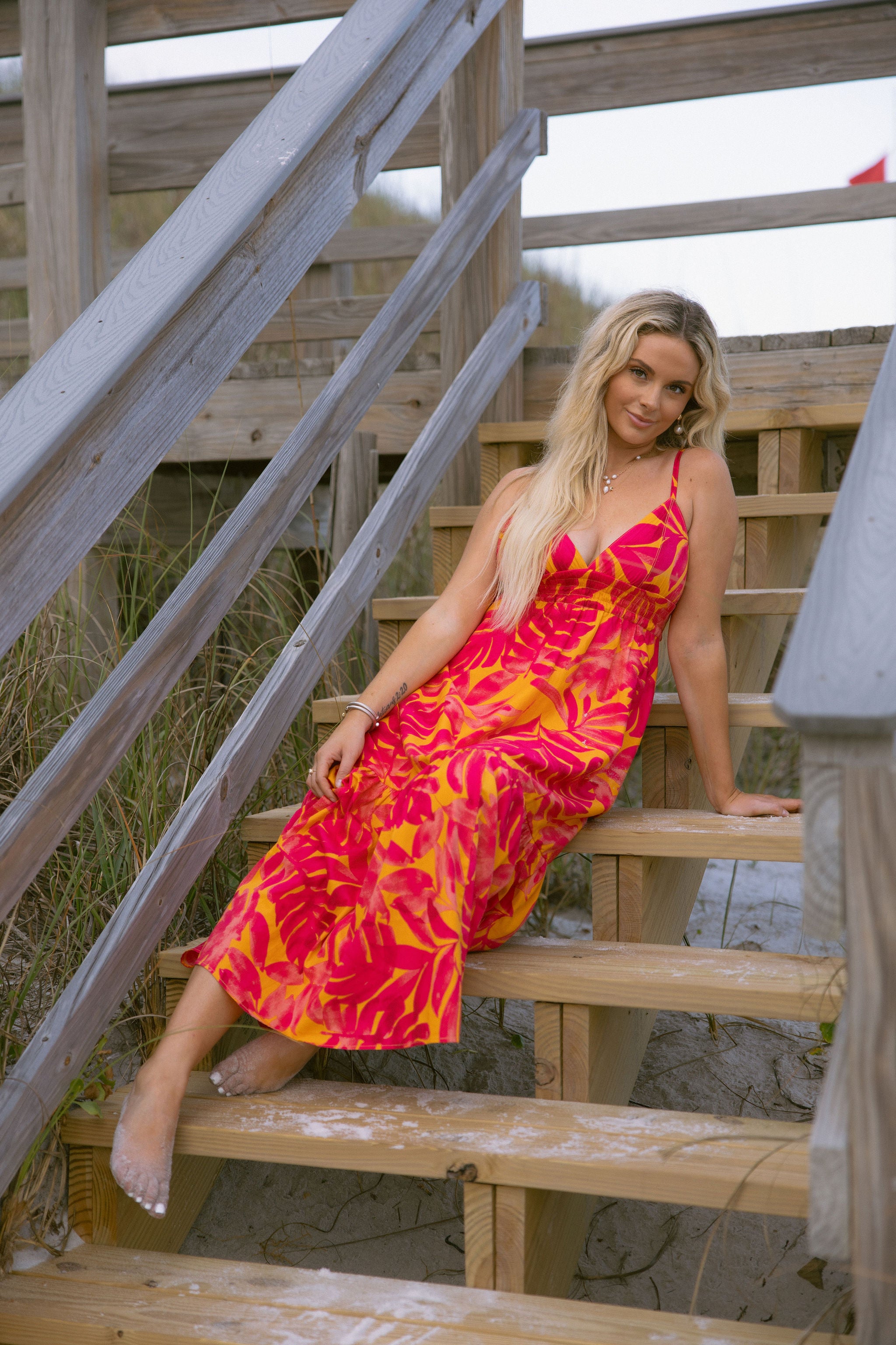 Full body front view of female model wearing the Melina Orange & Pink Floral Maxi Dress that has bright orange and pink floral pattern, adjustable straps, and an open back. Model is laying on stairs.