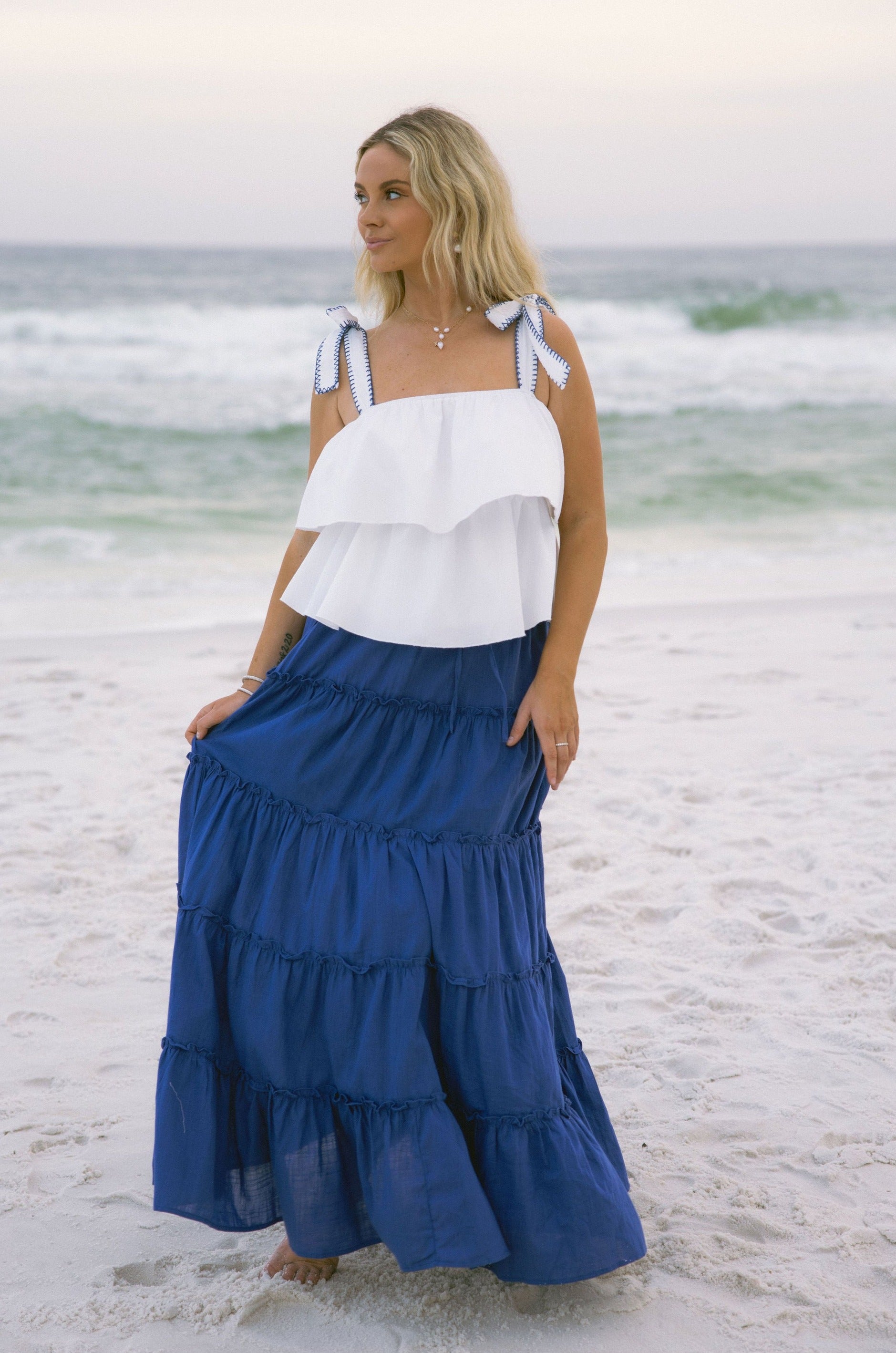 Full body front view of female model wearing the Danielle Blue Tiered Maxi Skirt that has royal blue fabric, a tiered body, and elastic waist. Worn with white tank top.