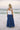 Full body back view of female model wearing the Madelyn White Tie Strap Tank Top that has tiered white fabric and tie straps with navy trim. Worn with blue skirt.