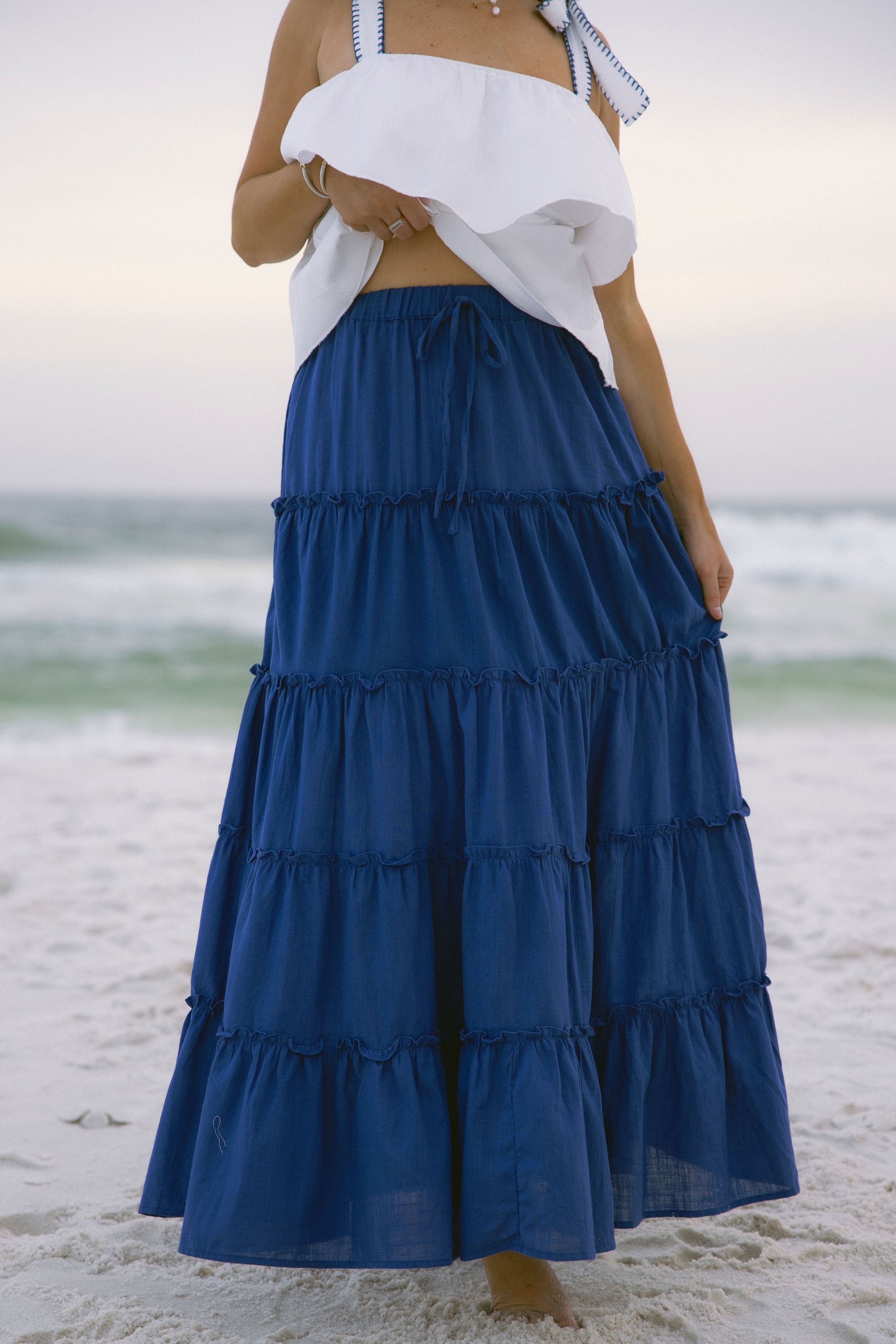 Lower body front view of female model wearing the Danielle Blue Tiered Maxi Skirt that has royal blue fabric, a tiered body, and elastic waist. Worn with white tank top.