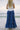 Lower body back view of female model wearing the Danielle Blue Tiered Maxi Skirt that has royal blue fabric, a tiered body, and elastic waist. Worn with white tank top.