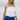 Upper body front view of female model wearing the Madelyn White Tie Strap Tank Top that has tiered white fabric and tie straps with navy trim. Worn with blue skirt.