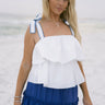 Upper body front view of female model wearing the Madelyn White Tie Strap Tank Top that has tiered white fabric and tie straps with navy trim. Worn with blue skirt.
