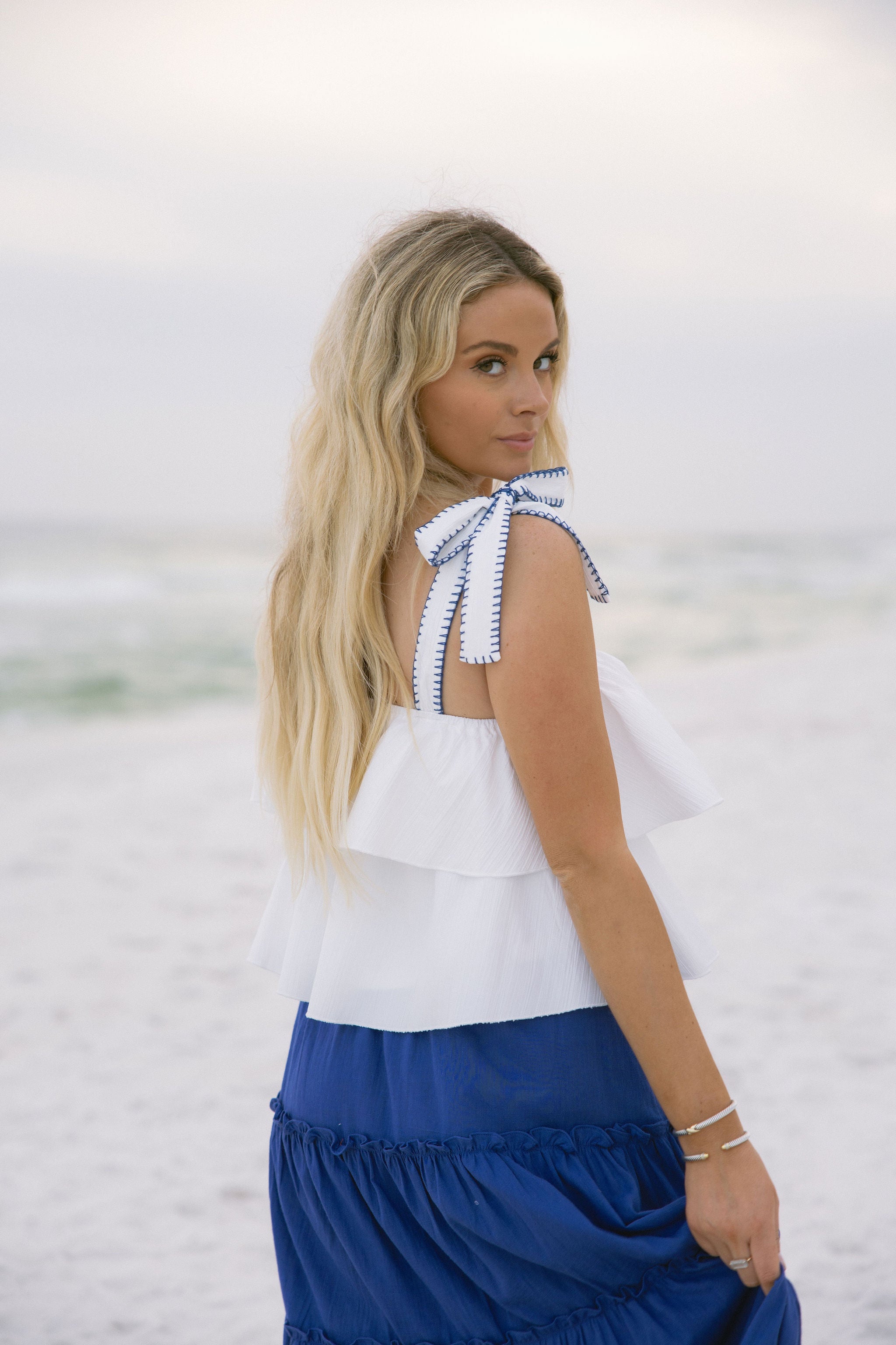 Upper body side view of female model wearing the Madelyn White Tie Strap Tank Top that has tiered white fabric and tie straps with navy trim. Worn with blue skirt.