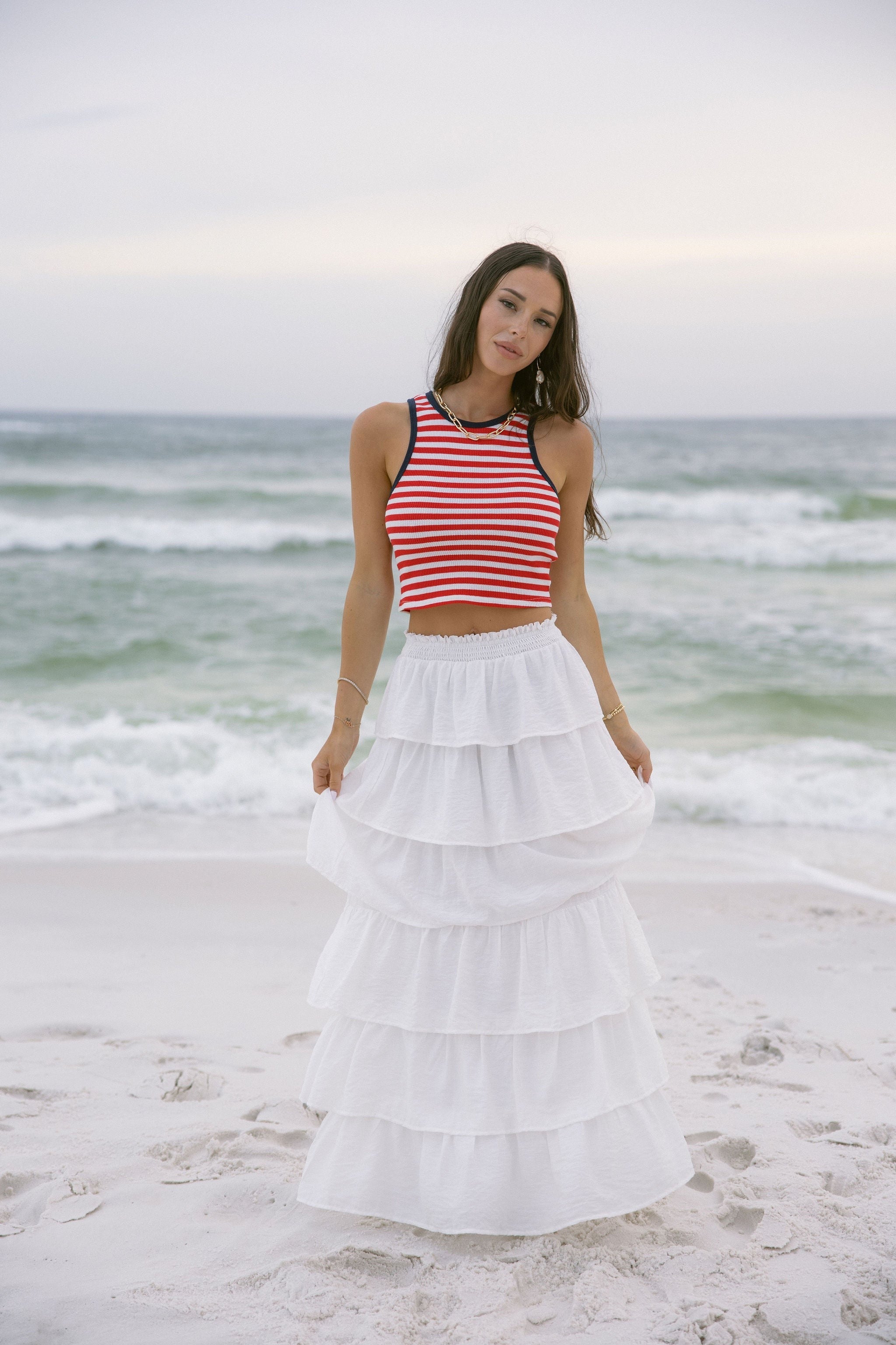 Upper body front view of female model wearing the Felicity Red, White, & Blue Striped Tank Top that has red and white horizontal stripes, navy trim, a cropped waist, and thick straps. Worn with white skirt.