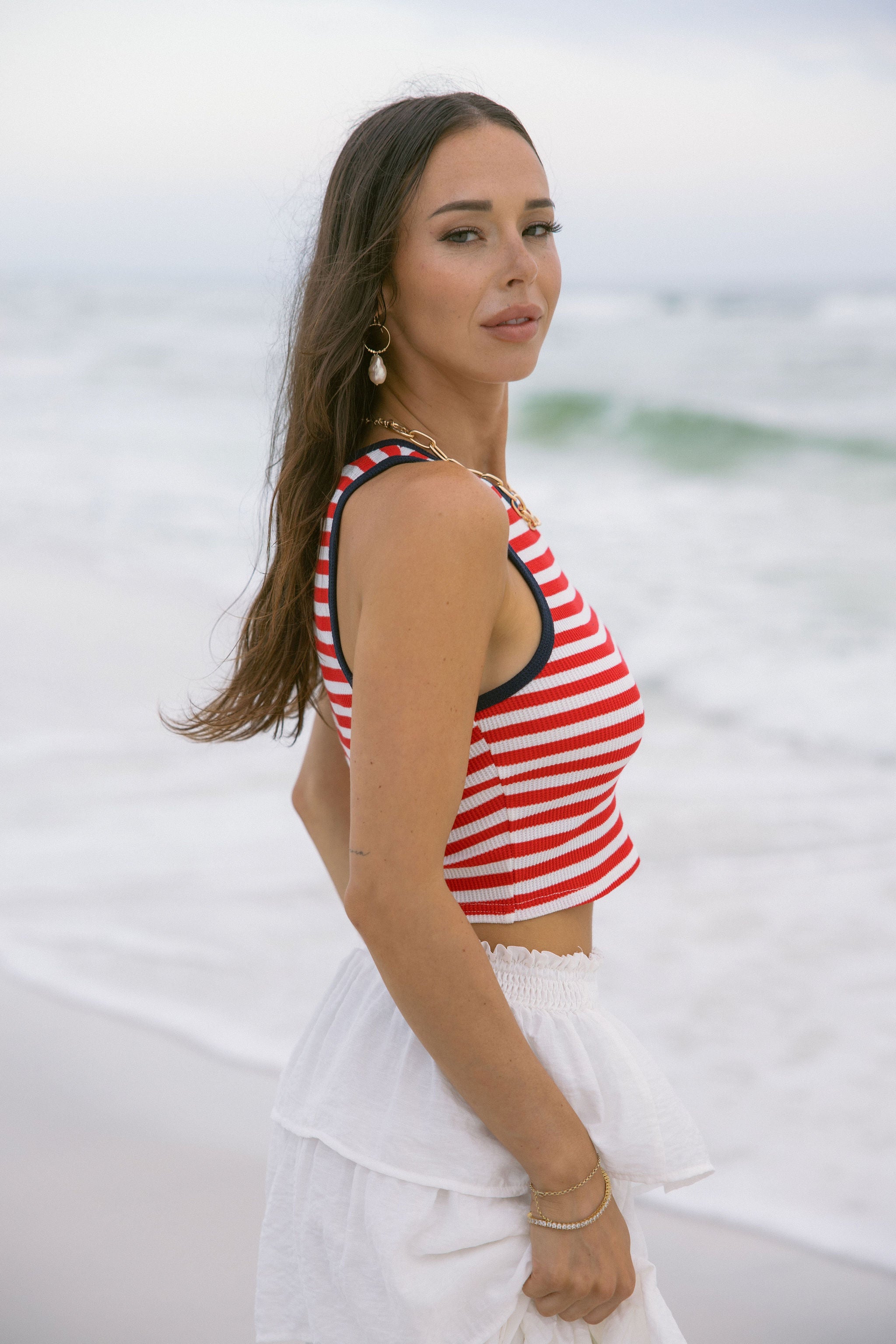 Upper body side view of female model wearing the Felicity Red, White, & Blue Striped Tank Top that has red and white horizontal stripes, navy trim, a cropped waist, and thick straps. Worn with white skirt.