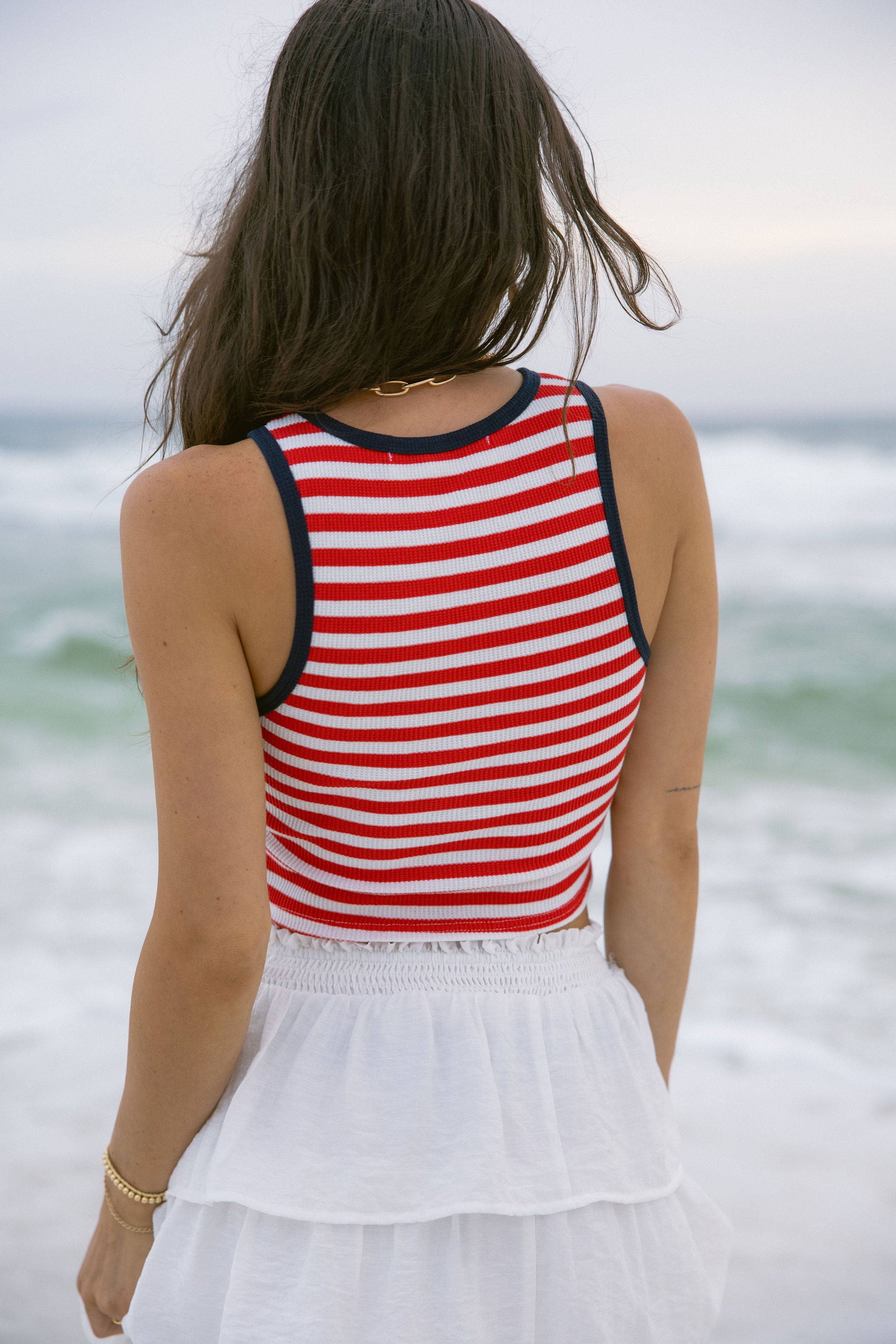 Upper body back view of female model wearing the Felicity Red, White, & Blue Striped Tank Top that has red and white horizontal stripes, navy trim, a cropped waist, and thick straps. Worn with white skirt.
