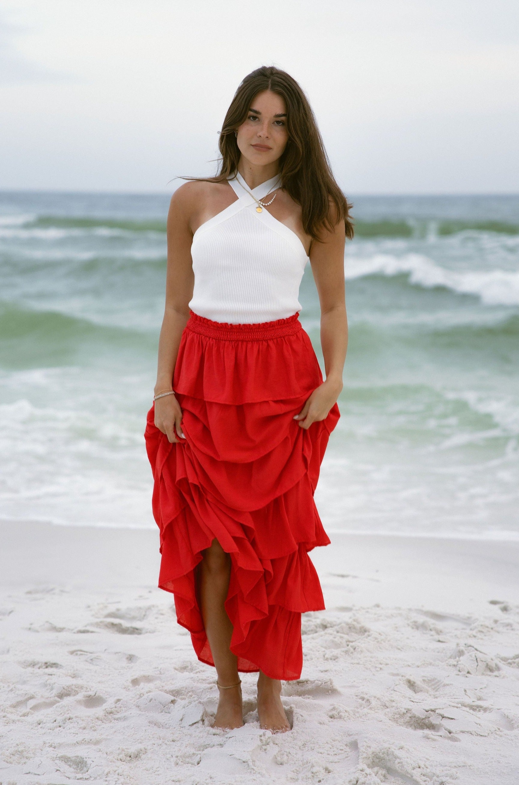 Full body front view of female model wearing the Katrina Tiered Ruffle Maxi Skirt in Red that has tiered red fabric and an elastic waist. Worn with white top tucked in. Model is holding skirt up.
