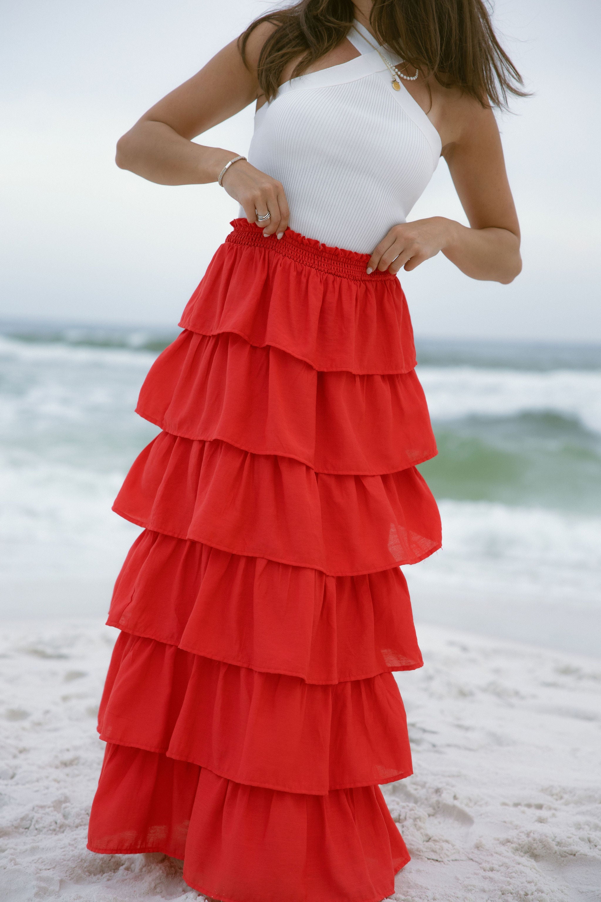 Lower body front view of female model wearing the Katrina Tiered Ruffle Maxi Skirt in Red that has tiered red fabric and an elastic waist. Worn with white top tucked in.
