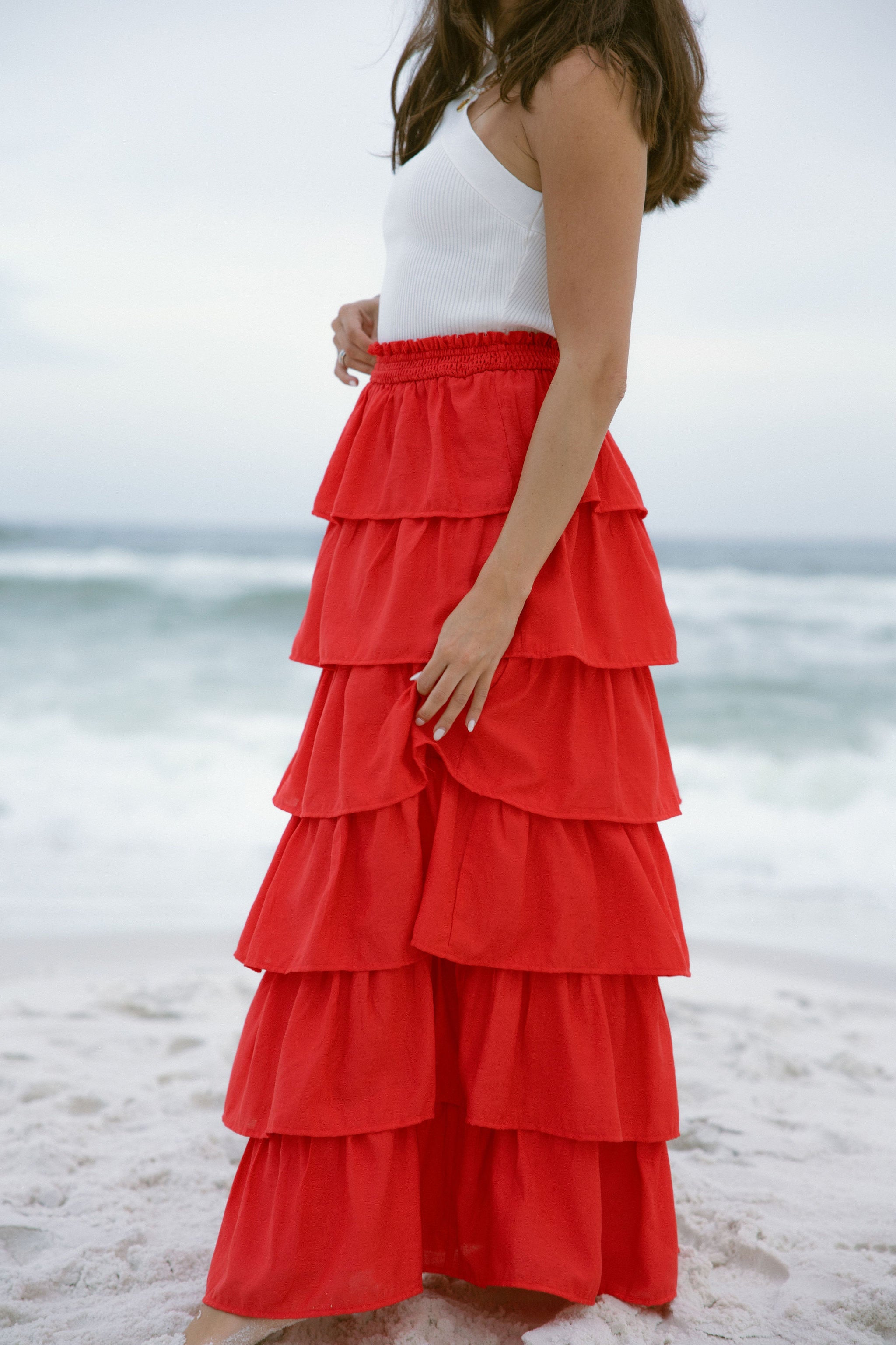 Lower body side view of female model wearing the Katrina Tiered Ruffle Maxi Skirt in Red that has tiered red fabric and an elastic waist. Worn with white top tucked in.