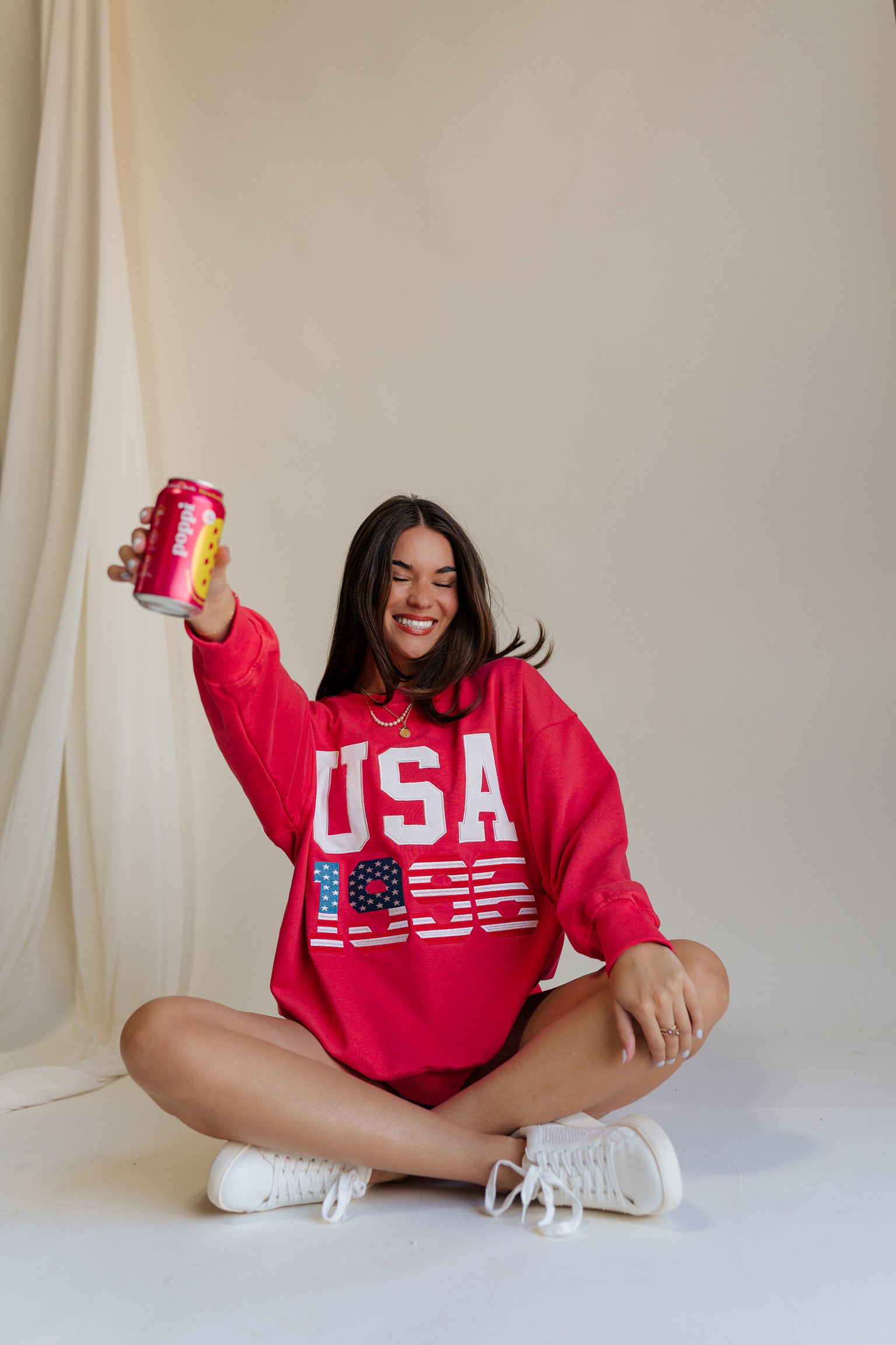 front view of female model sitting on the floor wearing the USA 1996 Red Long Sleeve Sweatshirt which features Lightweight Red Fabric, Round Neckline, Long Sleeves, Ribbed Hem and USA 1996 graphic with white, red and blue stitch design