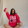 front view of female model sitting on the floor wearing the USA 1996 Red Long Sleeve Sweatshirt which features Lightweight Red Fabric, Round Neckline, Long Sleeves, Ribbed Hem and USA 1996 graphic with white, red and blue stitch design