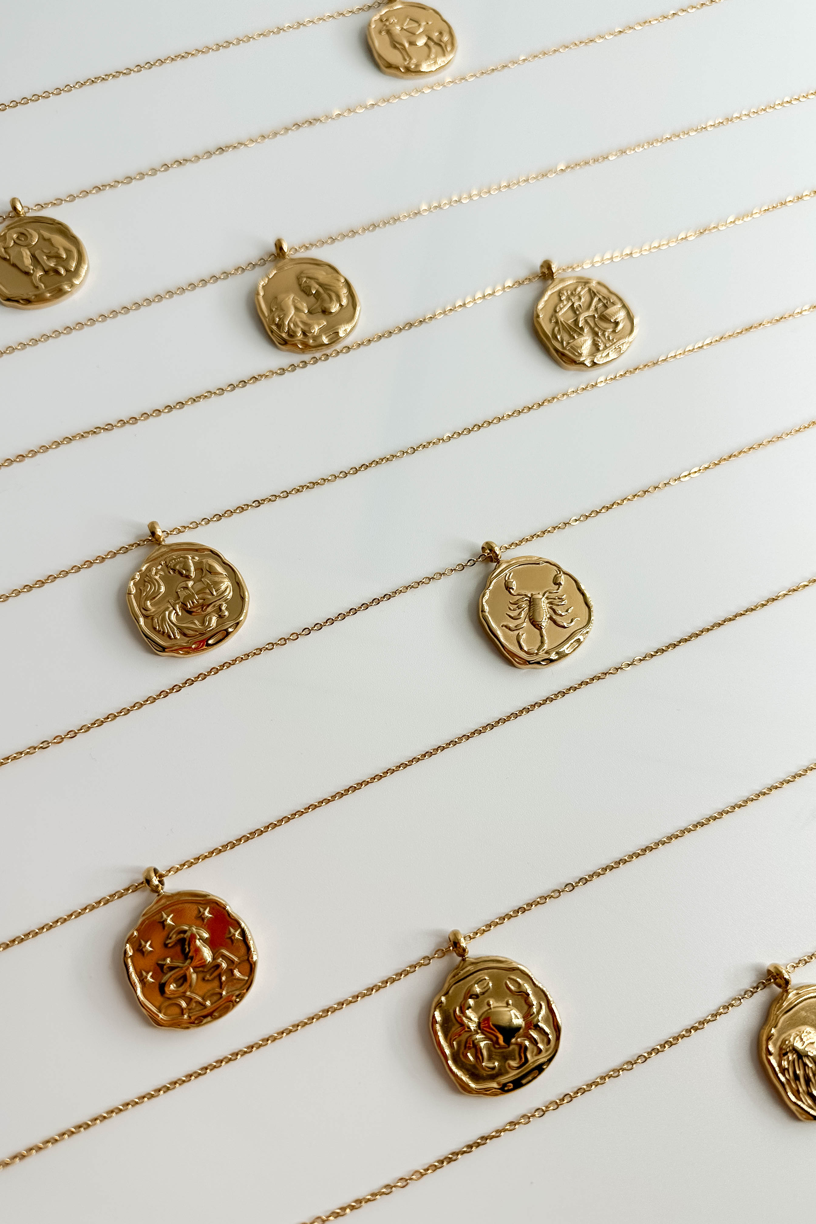 Image shows 9 of the Zodiac Gold Coin Necklaces lined up against a white background. 