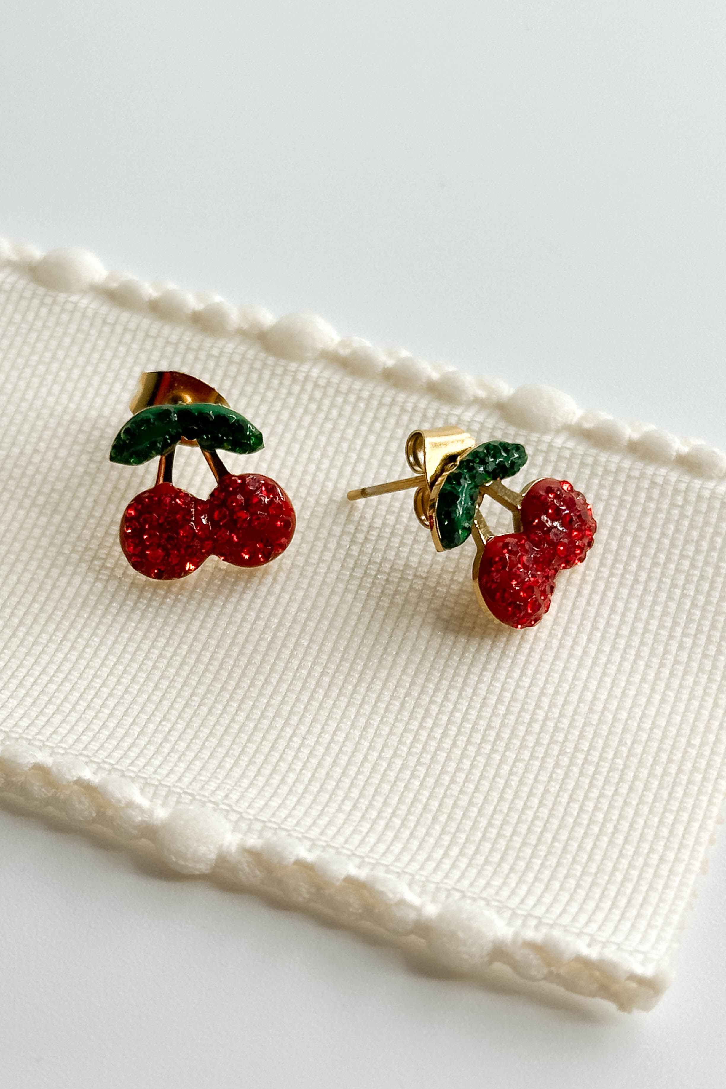 Image shows the Penelope Cherry Stud Earrings against a white background. Each earring has two red cherries with two green leaves, made from rhinestones on stud background