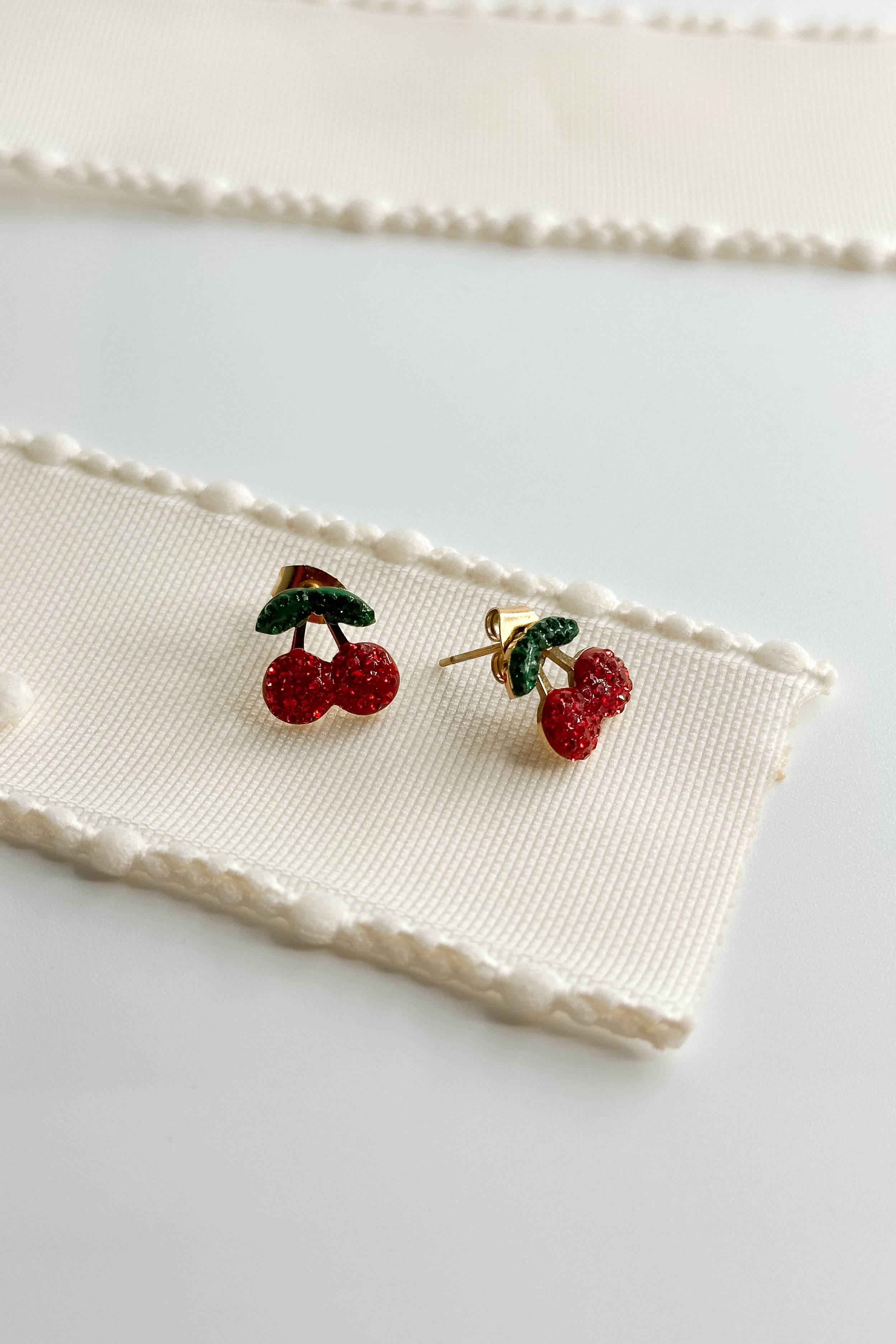 Image shows the Penelope Cherry Stud Earrings against a white background. Each earring has two red cherries with two green leaves, made from rhinestones on stud background