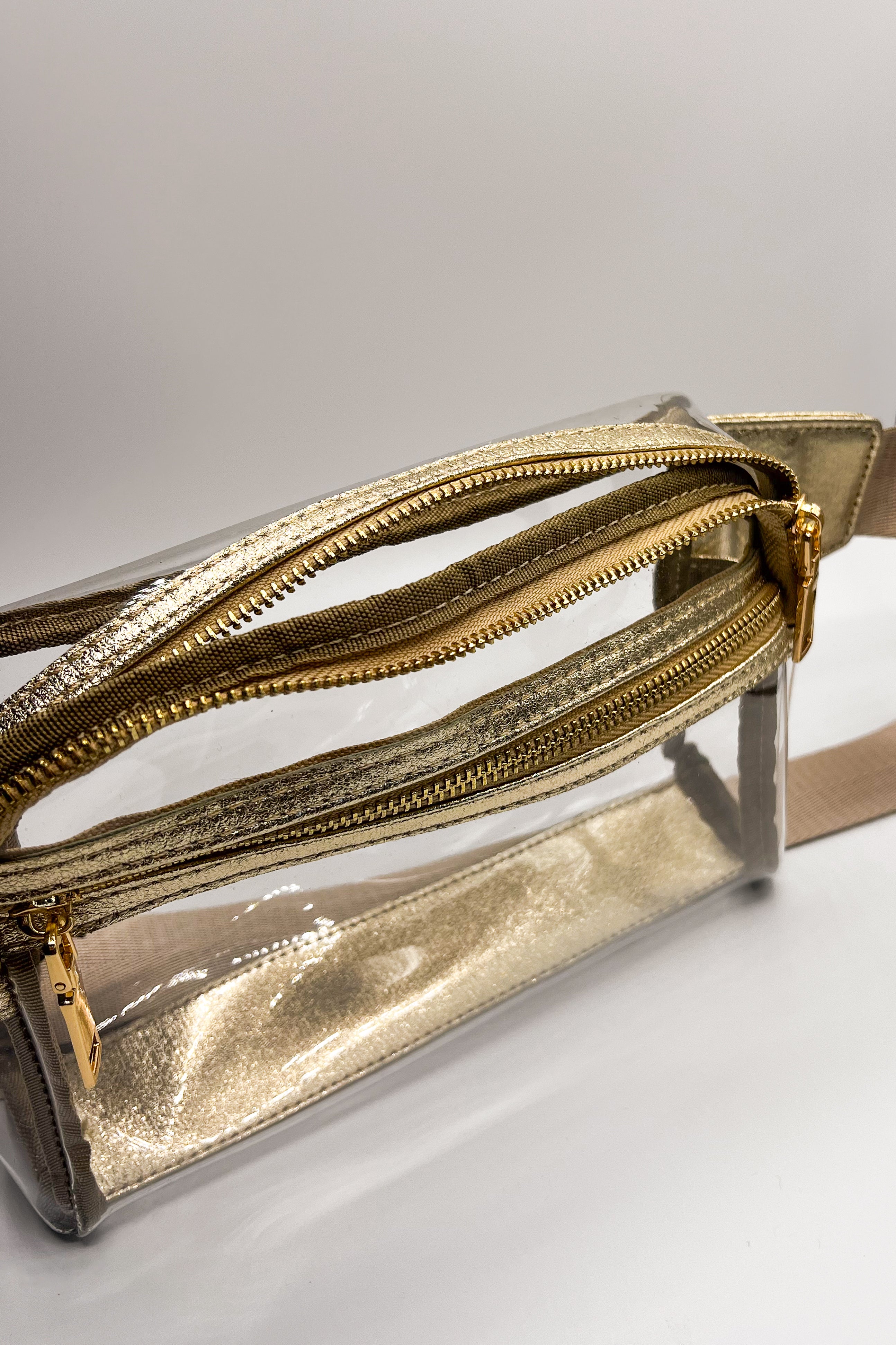 Close up of the Parker Clear and Gold Sling Bag. This features clear fabric with gold trim, two pockets with gold zipper closure and pink adjustable strap with gold buckle hardware.