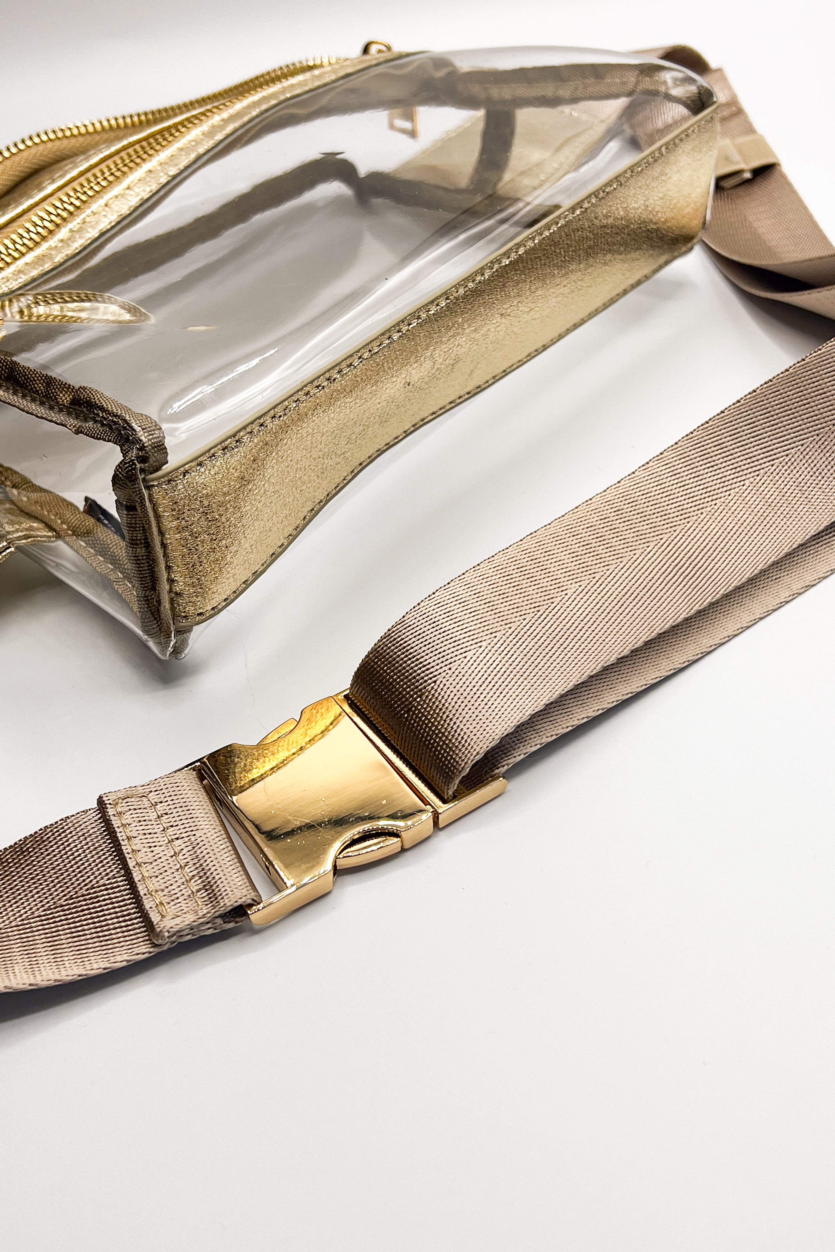 Close up of the Parker Clear and Gold Sling Bag. This features clear fabric with gold trim, two pockets with gold zipper closure and pink adjustable strap with gold buckle hardware.