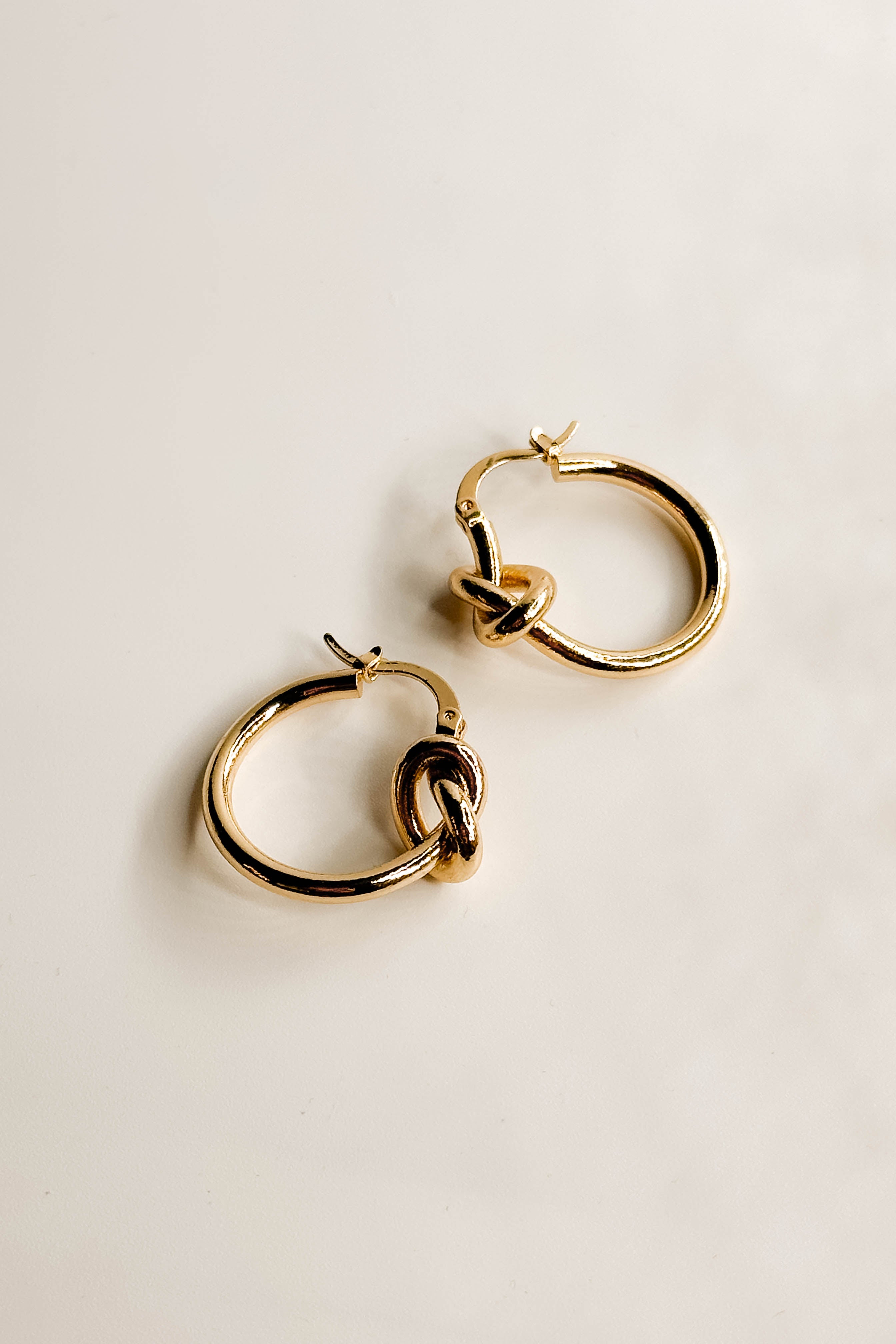 Close up side image of the Ava Gold Knot Hoop Earrings that feature small gold hoops with a knot design and clasp closure, shown on cream paper..
