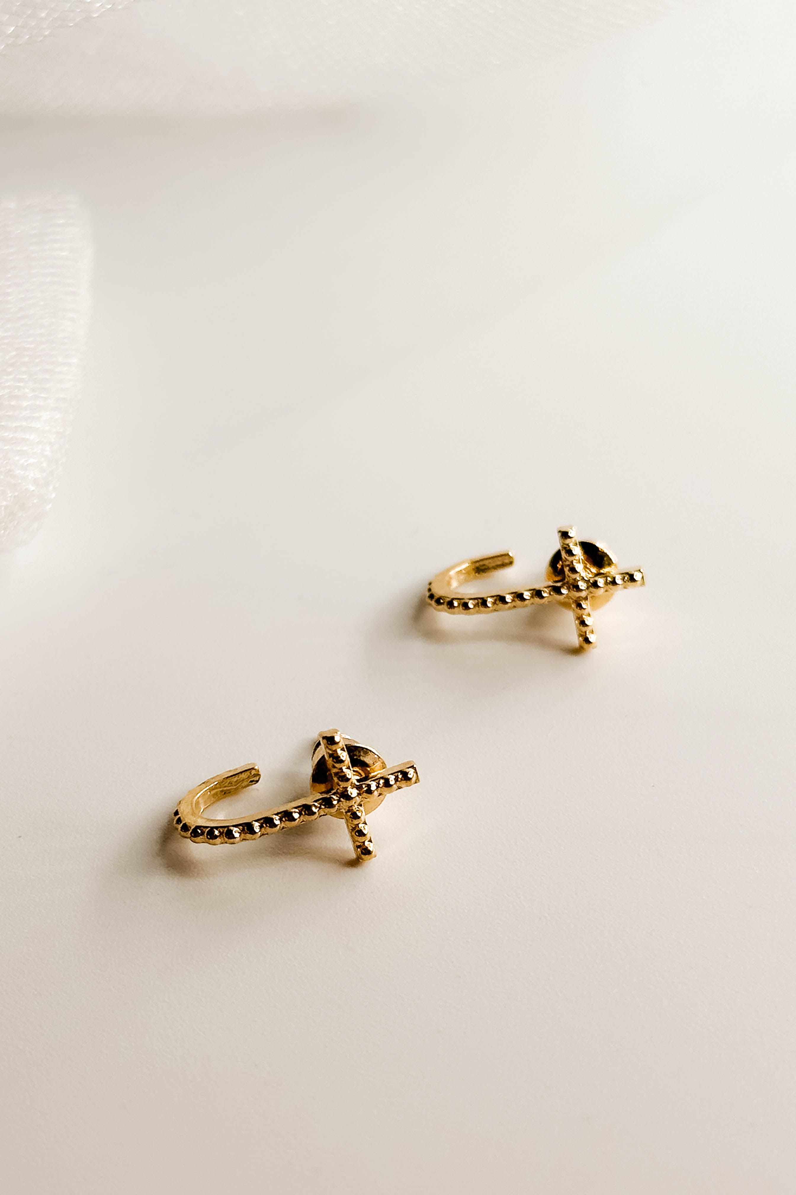 Close up detailed flat lay of the Grace Gold Cross Stud Earring which features mini gold, open cross shaped huggies with hobnail design.