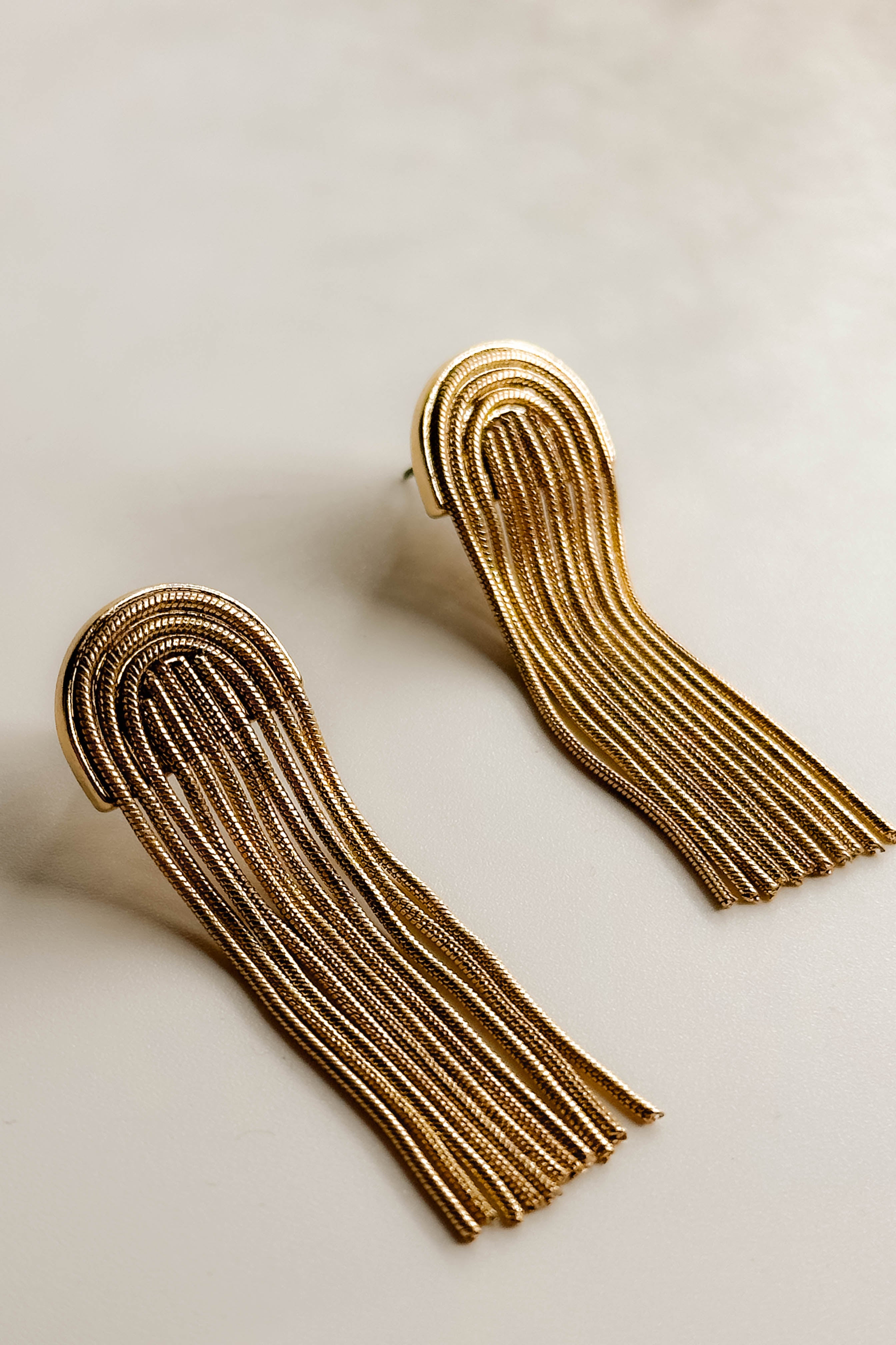 Top view of the Celine Gold Arch Fringe Earrings that feature gold arches on post backs with gold fringe, shown on white paper.