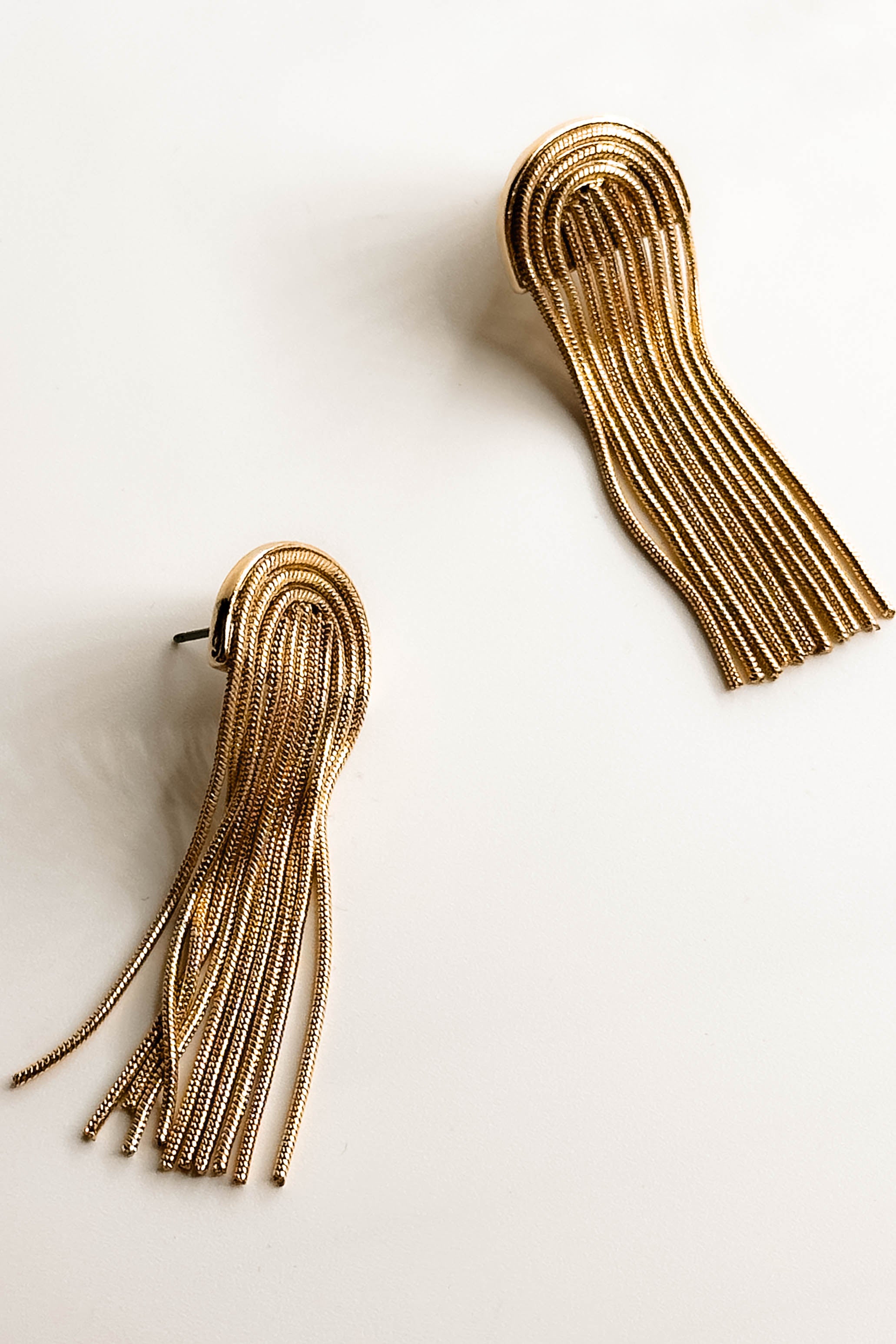 Top view of the Celine Gold Arch Fringe Earrings that feature gold arches on post backs with gold fringe, shown on white paper.