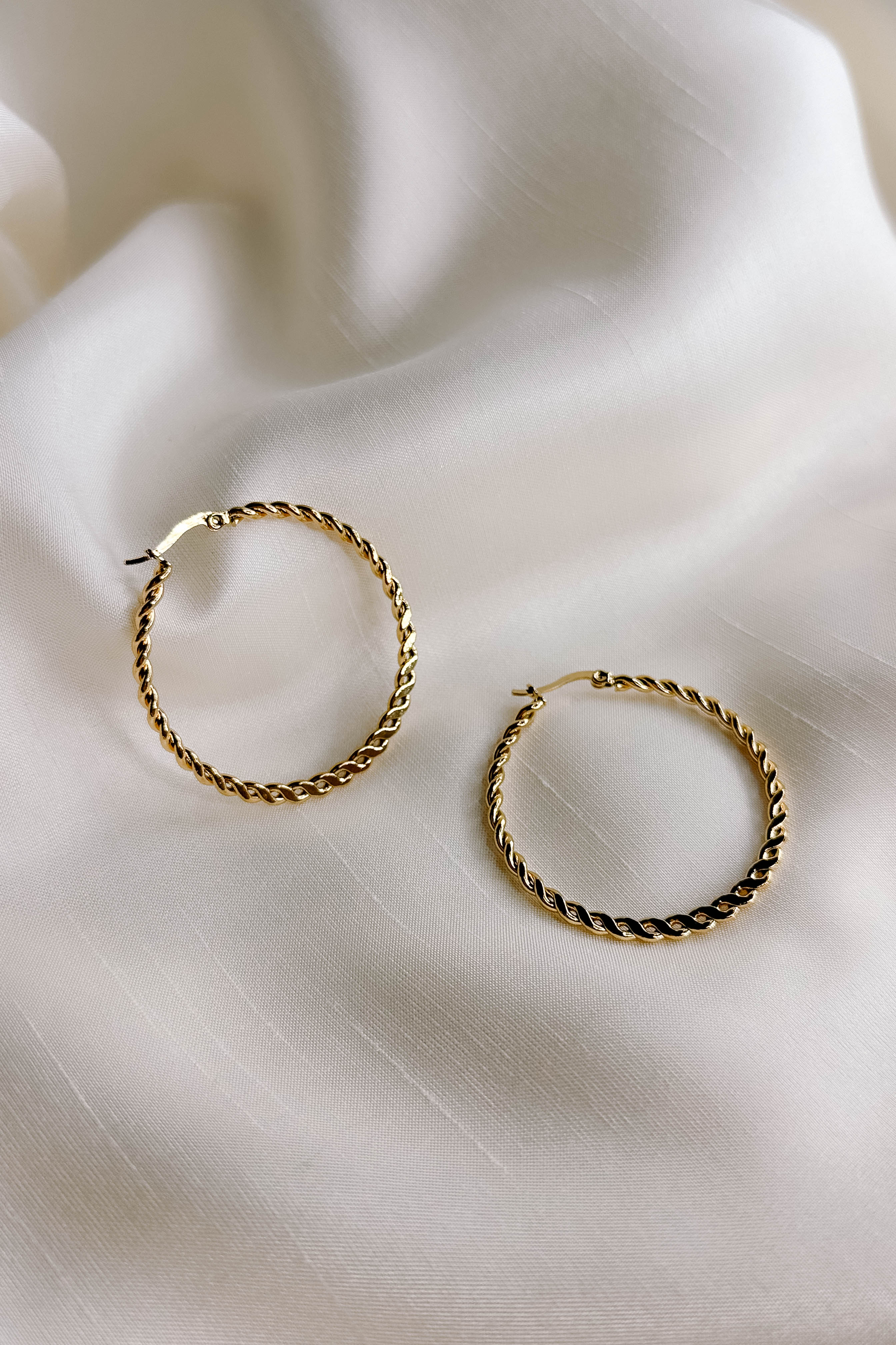 Top view of the Reese Gold Rope Hoop Earrings that feature medium closed hoops with a gold rope design and latch closures, shown against a cream fabric.