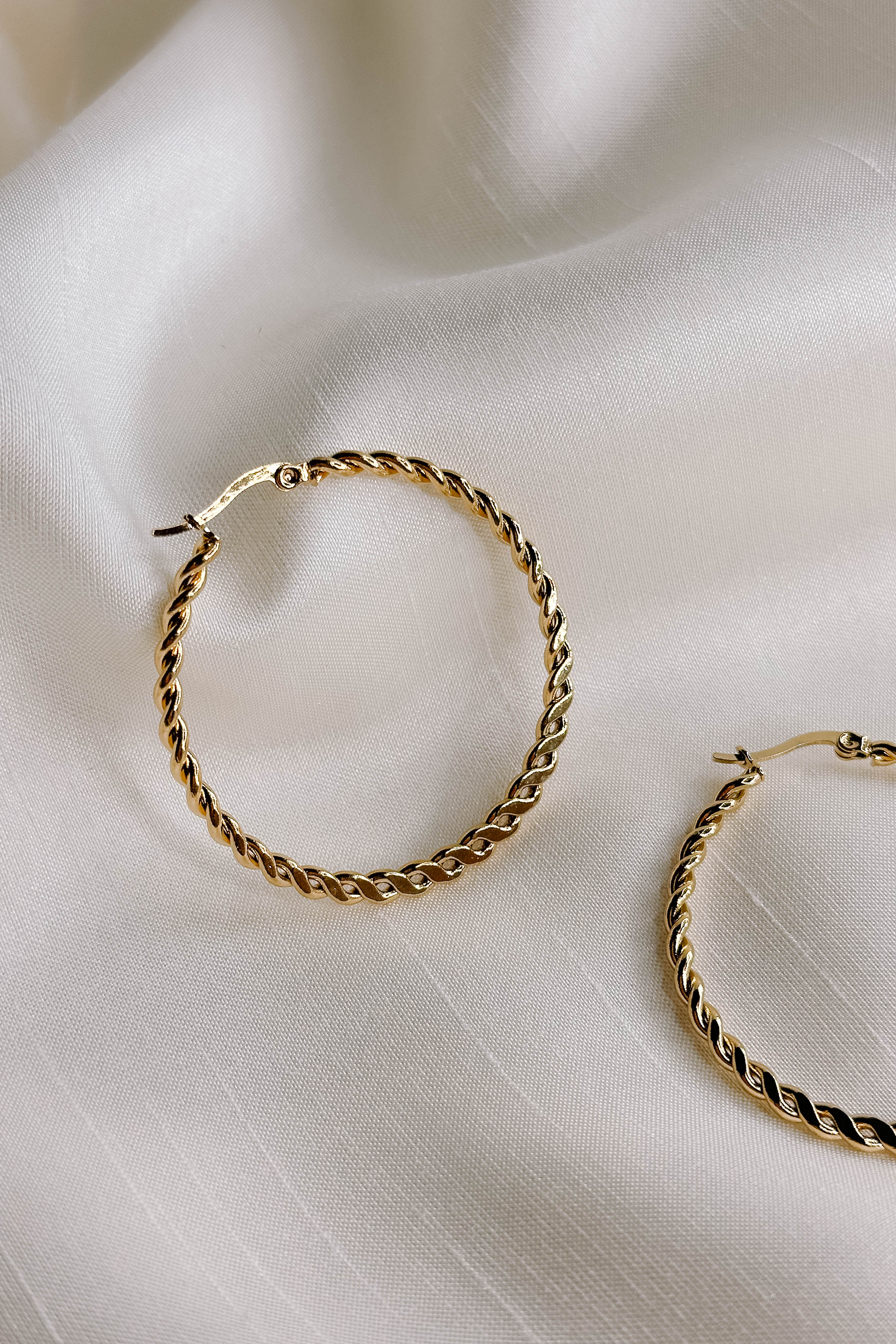 Side view of the Reese Gold Rope Hoop Earrings that feature medium closed hoops with a gold rope design and latch closures, shown against a cream fabric.