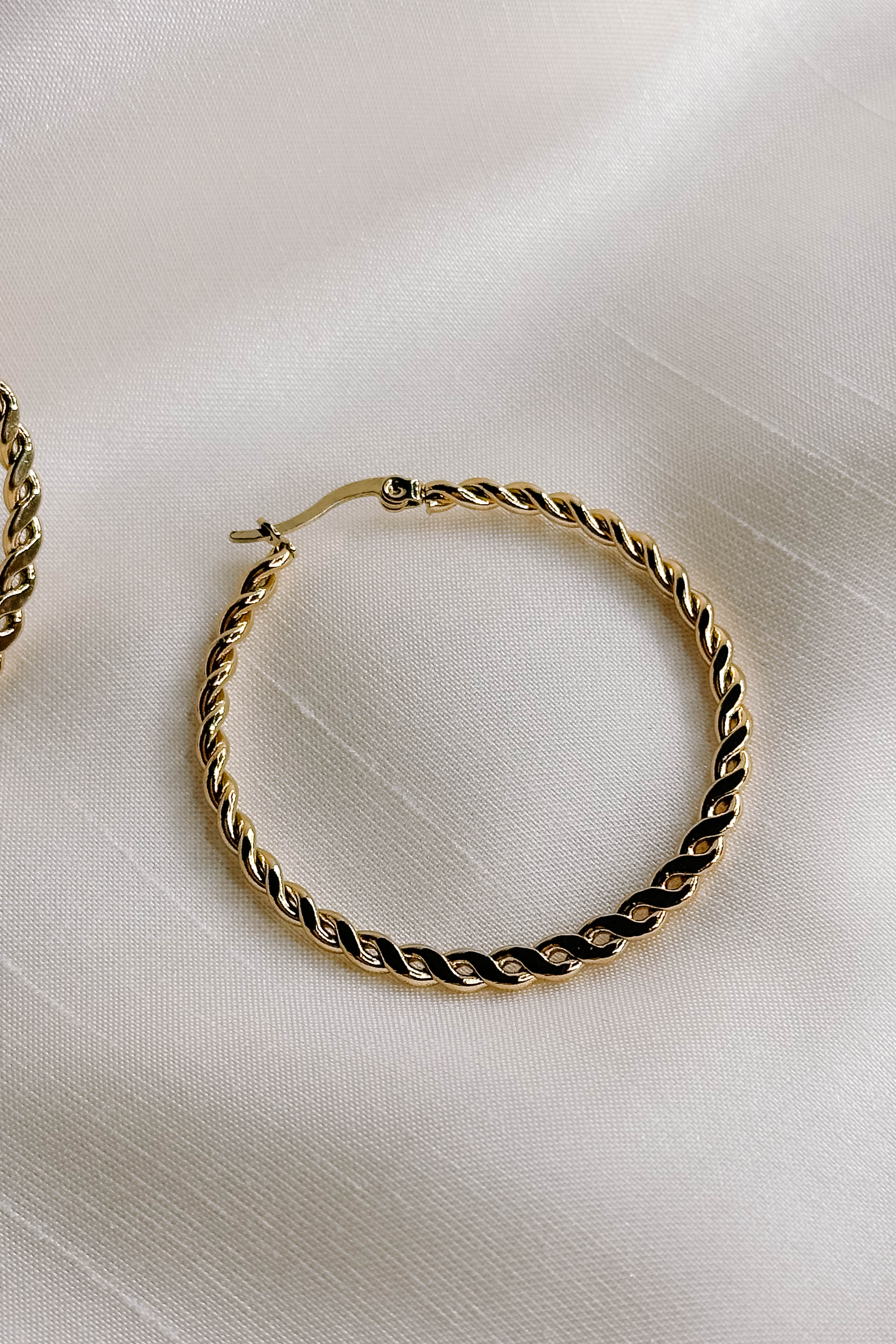 Side view of the Reese Gold Rope Hoop Earrings that feature medium closed hoops with a gold rope design and latch closures, shown against a cream fabric.