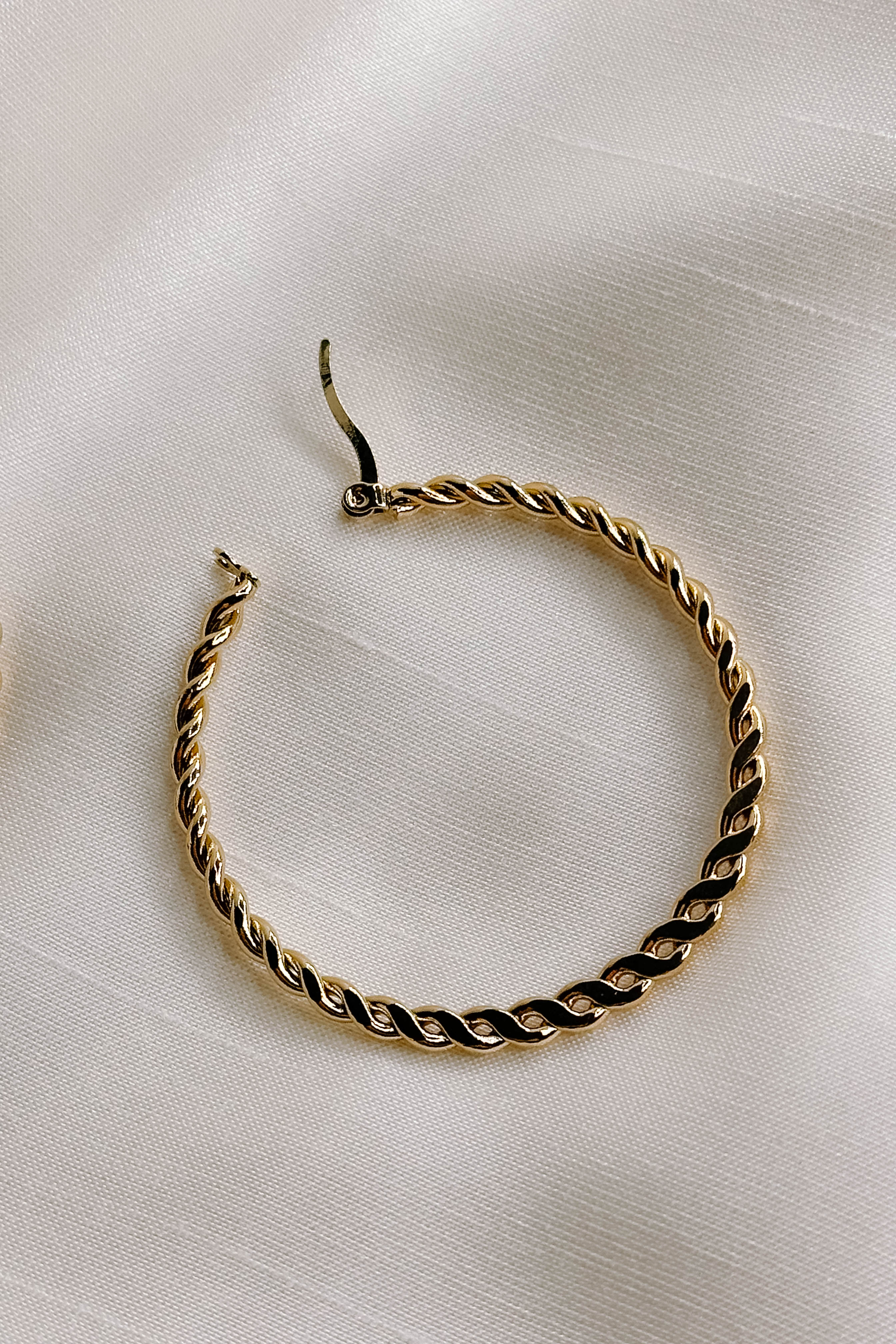 Single view of the Reese Gold Rope Hoop Earrings that feature medium closed hoops with a gold rope design and latch closures, shown against a cream fabric.