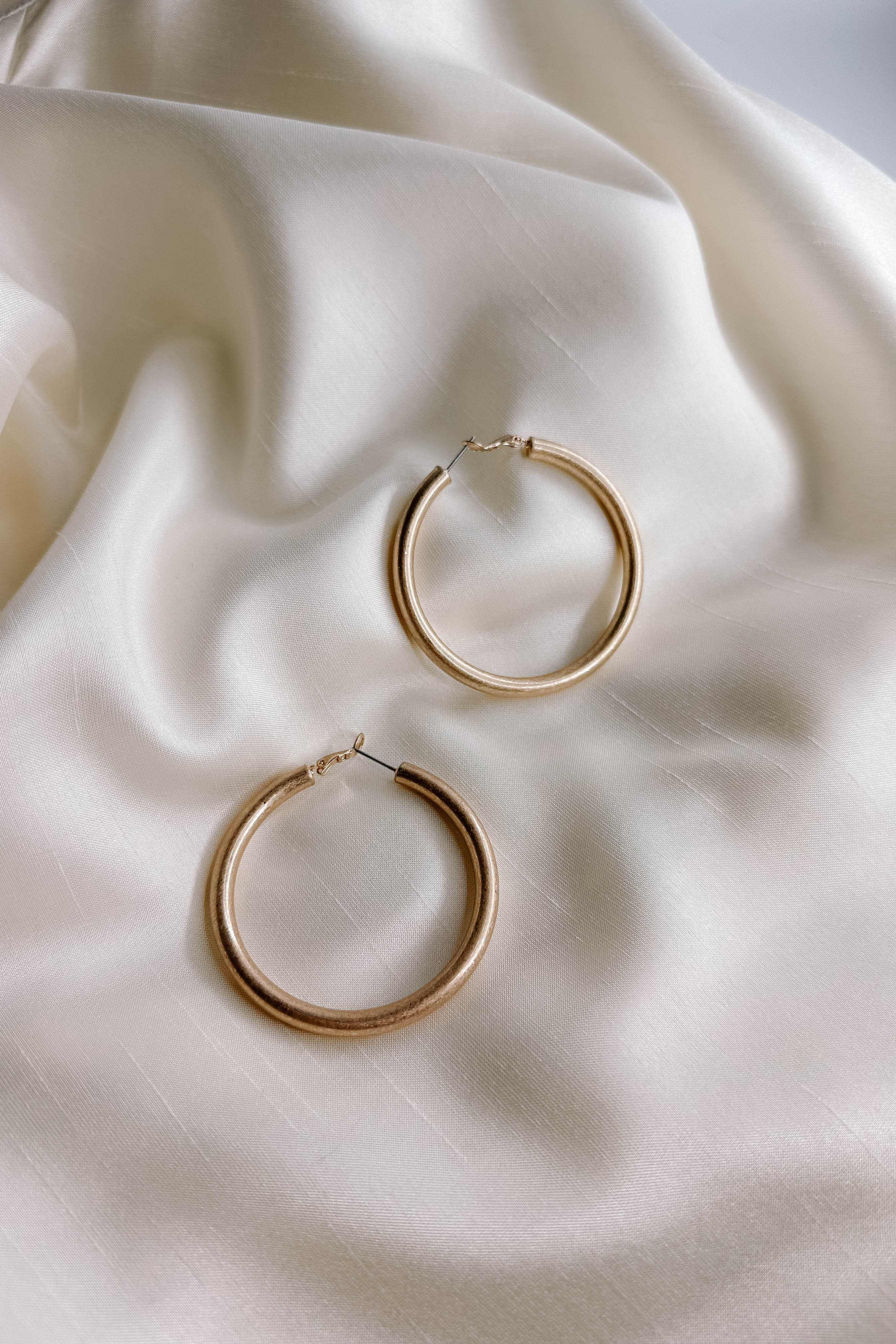 Close up top view of the Lucy Gold Brushed Hoop Earrings that features closed, medium brushed gold hoops with a hinged closure, shown on cream fabric.