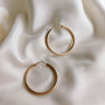 Close up top view of the Lucy Gold Brushed Hoop Earrings that features closed, medium brushed gold hoops with a hinged closure, shown on cream fabric.