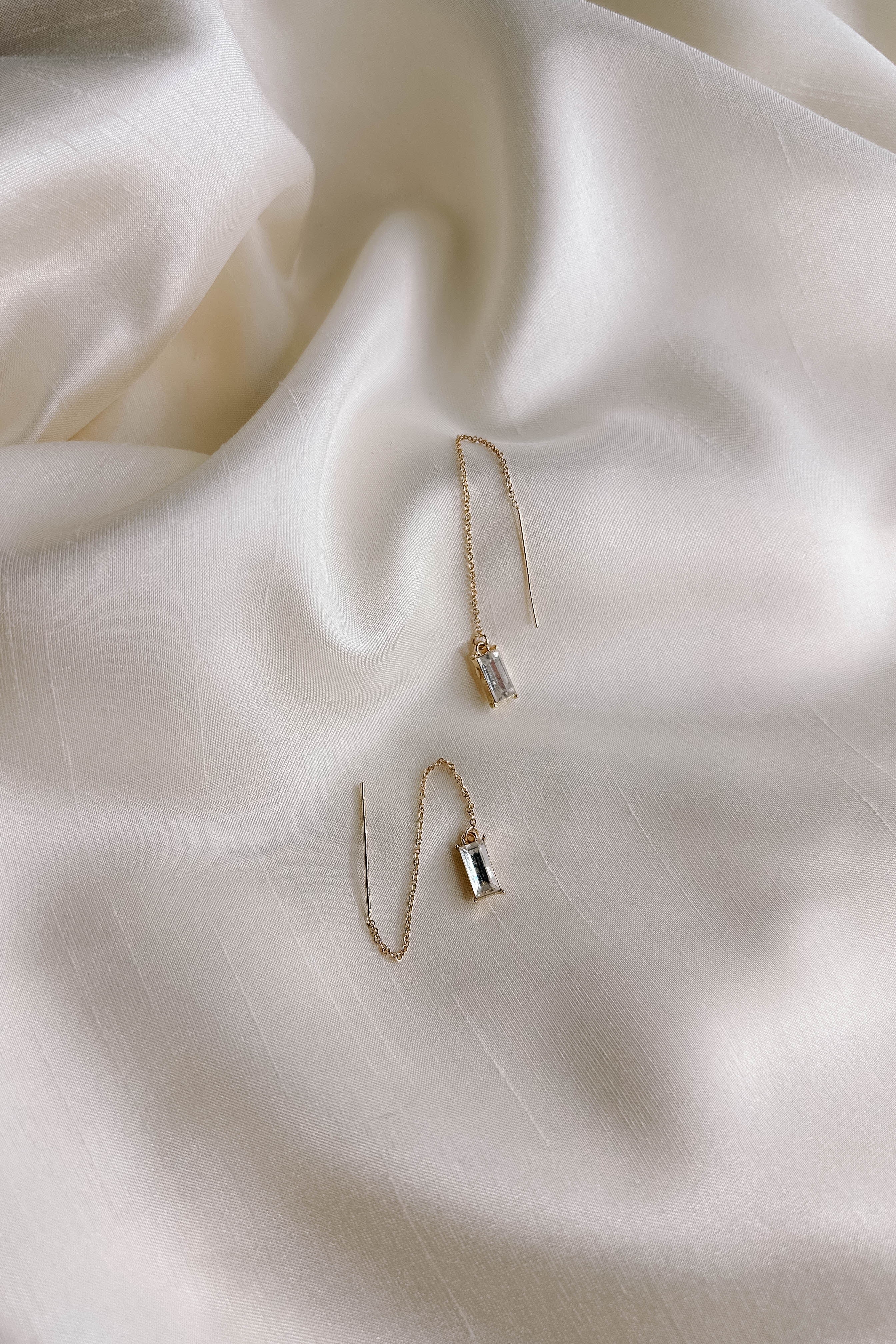 Top view of the Deena Gold Chain Rhinestone Dangle Earring which features gold dangle earrings linked to clear rectangle shaped stones, shown on cream fabric.
