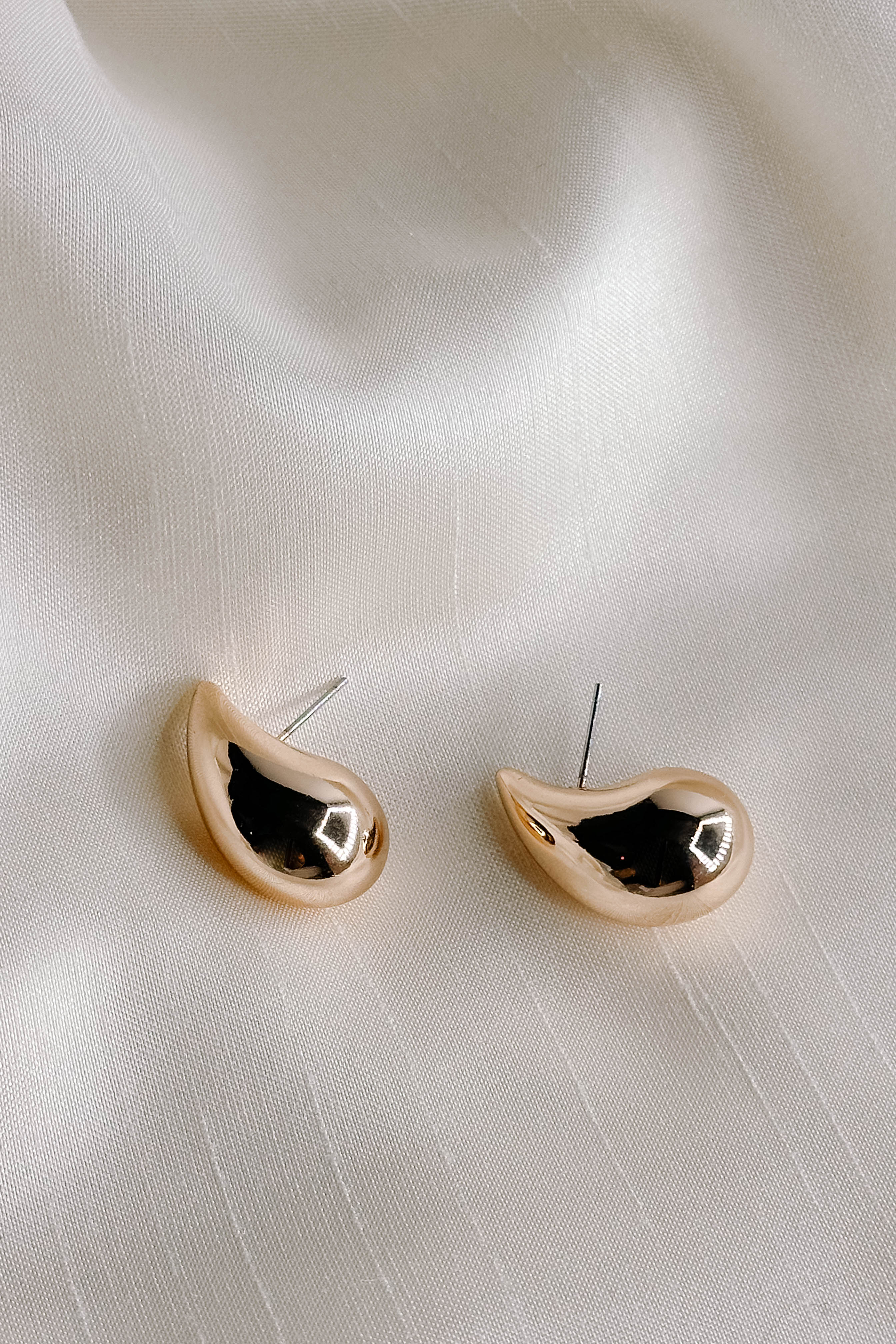 Side view flat lay of the Claire Gold Teardrop Stud Earrings that have thick gold scooped studs, shown on cream fabric..