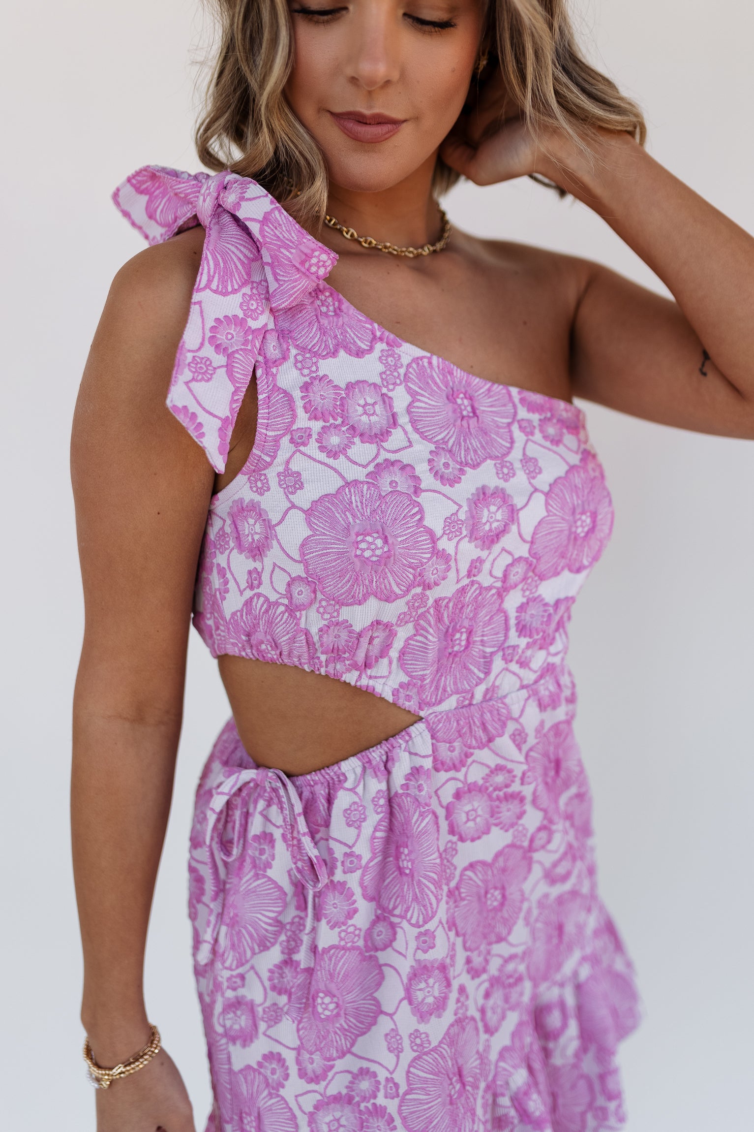 Close up view of model wearing the Laura Pink Floral One-Shoulder Dress which features light pink and pink textured fabric, mini length, flare hem skirt, floral print, elastic waistband, side cut-out detail with drawstring tie, one shoulder strap with bow detail and sleeveless.