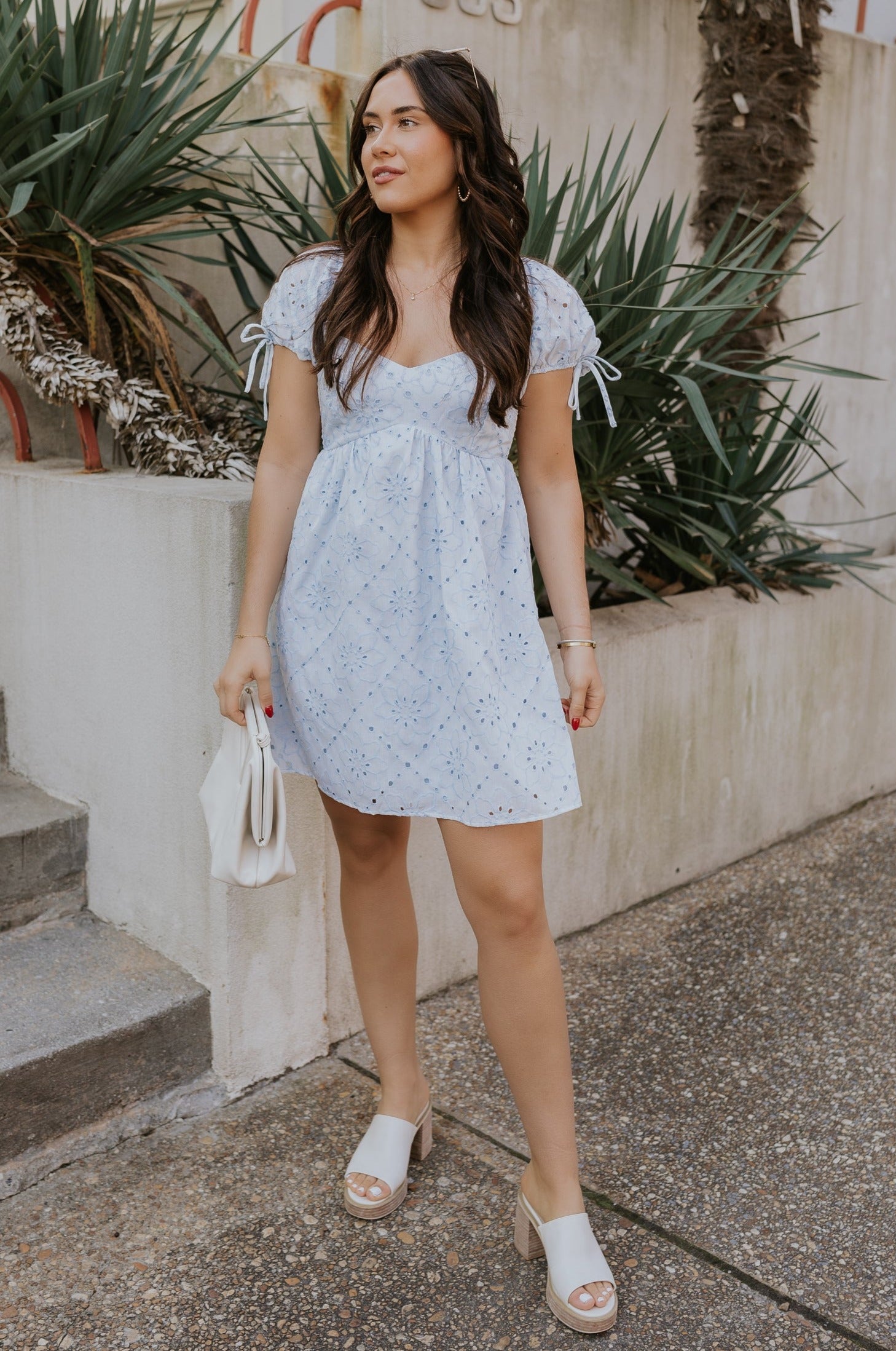 Full body view of female model wearing the The Ella Light Blue Eyelet Mini Dress which features light blue cotton fabric with an eyelet floral pattern, mini length, light blue lining, a sweetheart neckline, short puff sleeves with tie details, and a smocked back.