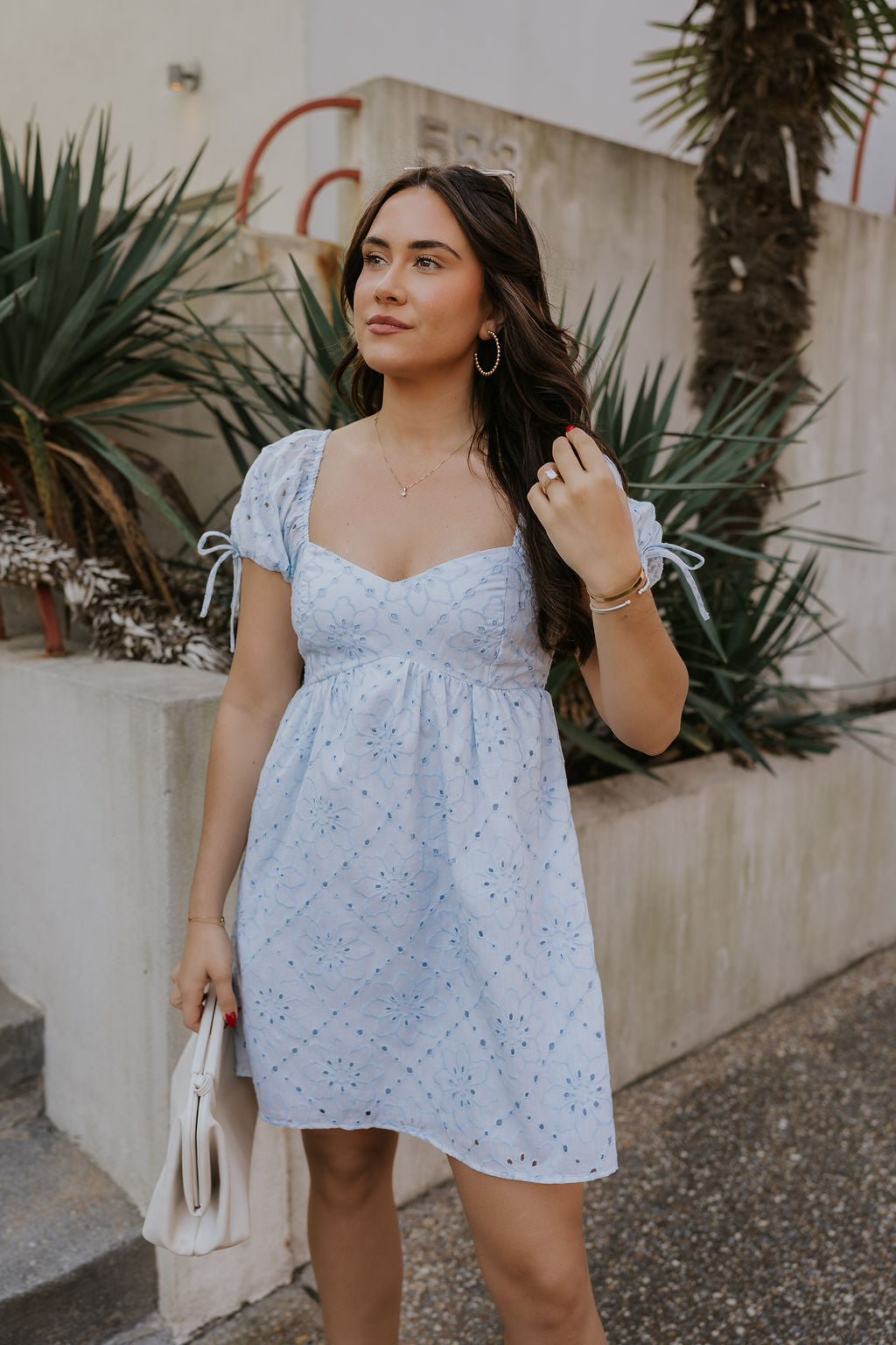 Front view of female model wearing the The Ella Light Blue Eyelet Mini Dress which features light blue cotton fabric with an eyelet floral pattern, mini length, light blue lining, a sweetheart neckline, short puff sleeves with tie details, and a smocked back.