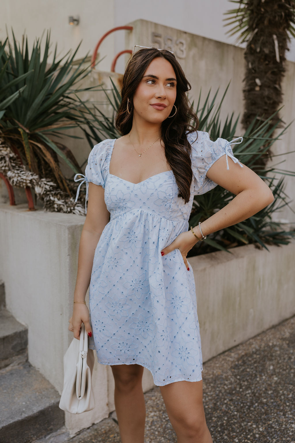 zfront view of female model wearing the The Ella Light Blue Eyelet Mini Dress which features light blue cotton fabric with an eyelet floral pattern, mini length, light blue lining, a sweetheart neckline, short puff sleeves with tie details, and a smocked back.